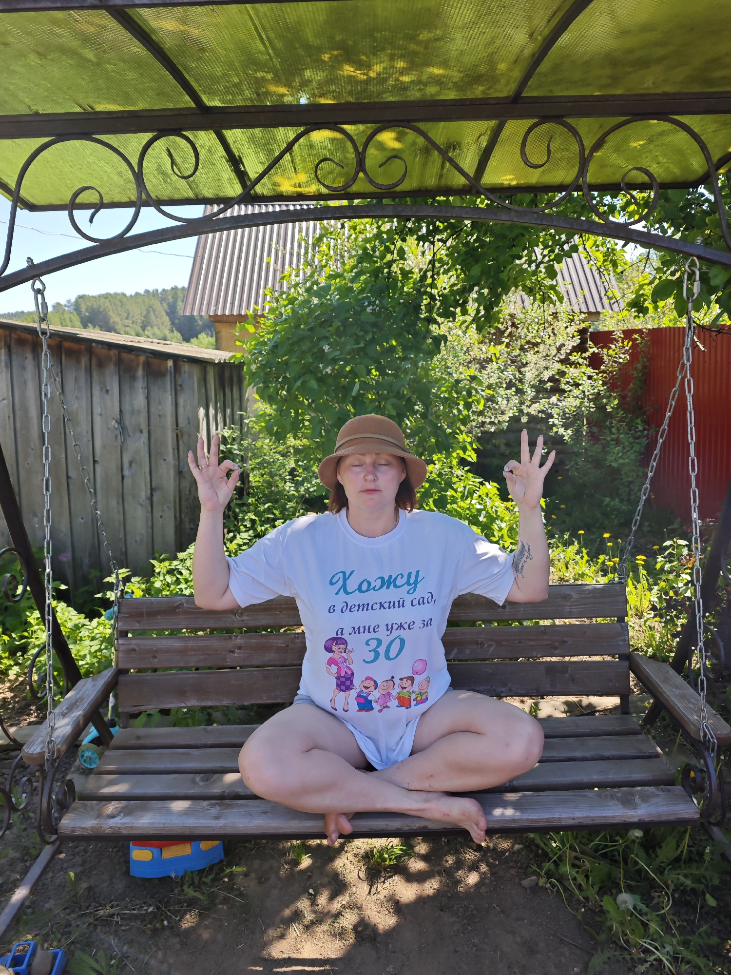 A woman sits cross-legged on a wooden swing, her arms raised in a meditative gesture. She wears a hat and a t-shirt with a graphic, and her eyes are closed. The swing is covered by a metal canopy with a translucent green roof, and it hangs in a yard with a wooden fence, various plants, and a red metal fence. The background includes a house with a metal roof and trees. The location is possibly Фоки, Russia.