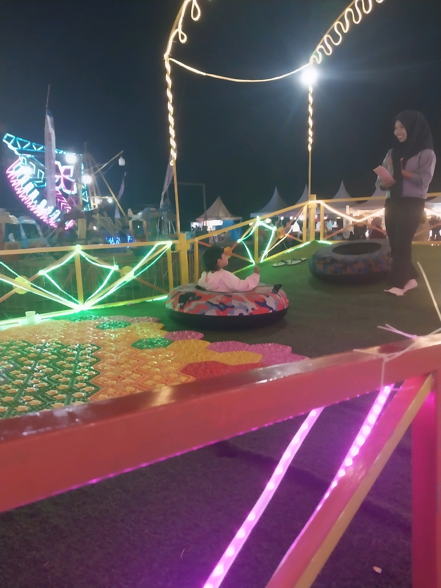 This is a night-time outdoor scene at a fairground in Jiwut, Indonesia. The focus is on a child sitting in a colorful inflatable tube. To the right, a woman wearing a hijab stands and appears to be holding a pink object. The ground is covered in artificial grass, and a vibrantly patterned mat is visible in the foreground. Behind the child, another inflatable tube lies on the ground. In the background, a Ferris wheel with colorful lights is partially visible, along with tents and other fairground attractions. The area is illuminated by various string lights, including yellow and green, as well as pink LED lights along the railing in the foreground. The overall atmosphere suggests a lively amusement park setting.