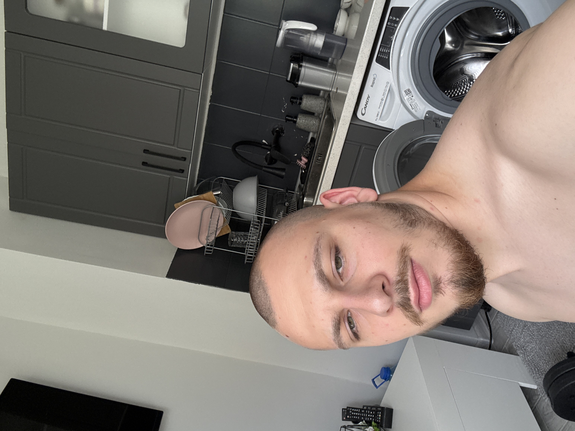 A close-up, slightly angled self-portrait of a man indoors. The man has a shaved head with short, dark hair, light skin, and a short beard. He is shirtless, revealing part of his torso.
The background consists of a kitchen area. Elements visible in the background include gray kitchen cabinets, a sink with various containers and a faucet, and a white washing machine. Dishes are drying on a rack.
The location is an apartment in Stara Zagora, Bulgaria.