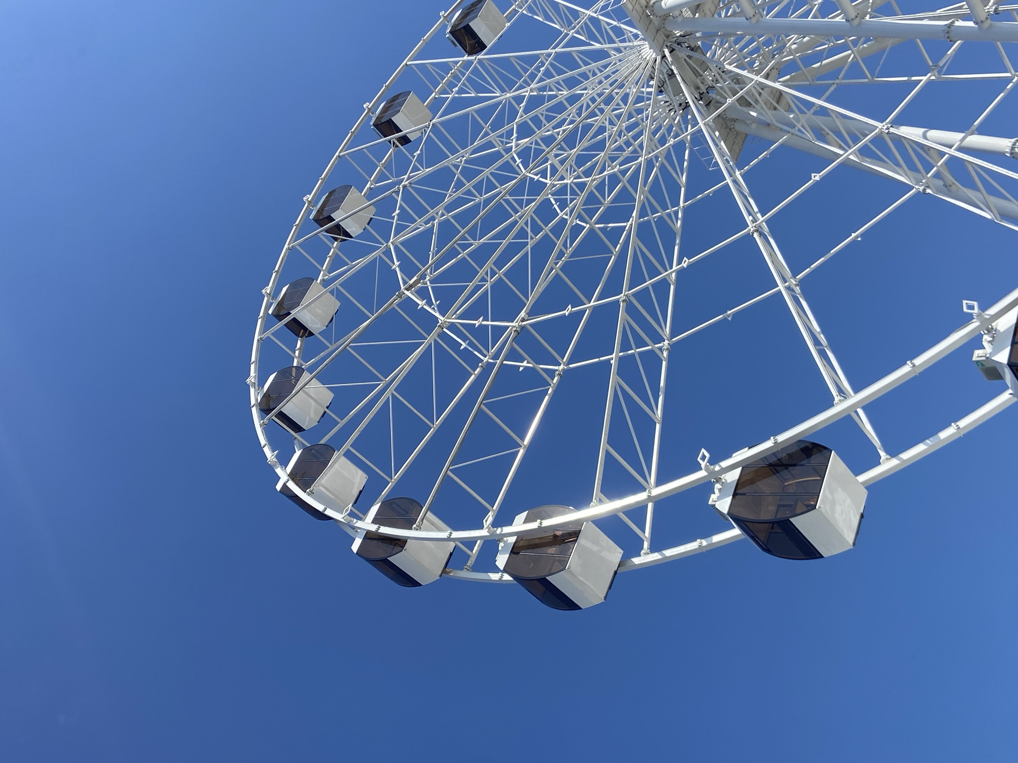 The image shows a low-angle view of a white Ferris wheel against a clear blue sky. The cabins of the wheel are boxy in shape, with white and dark-tinted surfaces. The structure of the wheel is made of white metal beams. There are no people visible in the image. The scene appears to be taken in Saratov, Russia, based on the style of the Ferris wheel.