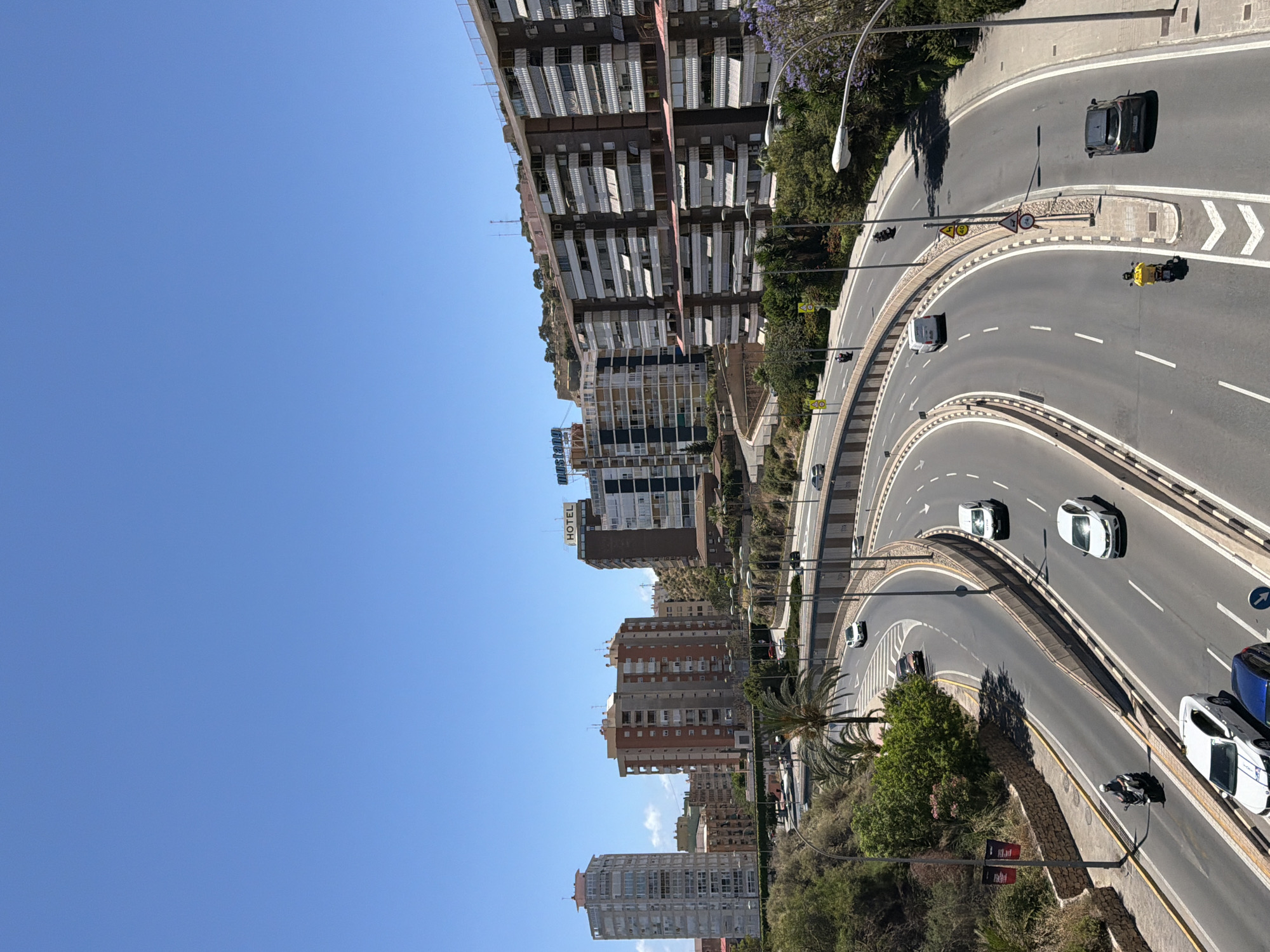 A high-angle, medium shot captures a wide, multi-lane highway curving through a city on a clear, bright day under a cloudless blue sky. On the left side of the highway, a steep, green, treed embankment rises, with a stone retaining wall visible near the bottom. A lamppost stands prominently on this side, adorned with two red banners. Several cars and a motorcycle are visible on the highway lanes.
Further up and to the left, a tall, slender, light-colored apartment building stands, with a pinkish-red top. To its right, a wider, reddish-brown apartment building with numerous balconies and white awnings is situated. In the background, more apartment buildings of varying heights and architectural styles dot the cityscape, some built into the hillside. One building features a prominent "HOTEL" sign.
The highway itself is paved and well-maintained, with clear white lane markings. Several vehicles, including cars, a van, and motorcycles, are in motion. One motorcycle carries a driver wearing a yellow backpack. The highway is bordered by concrete barriers and some landscaping, including palm trees and other greenery. In the distance, more buildings and hills are visible. The overall impression is one of urban activity and a sunny, warm climate.