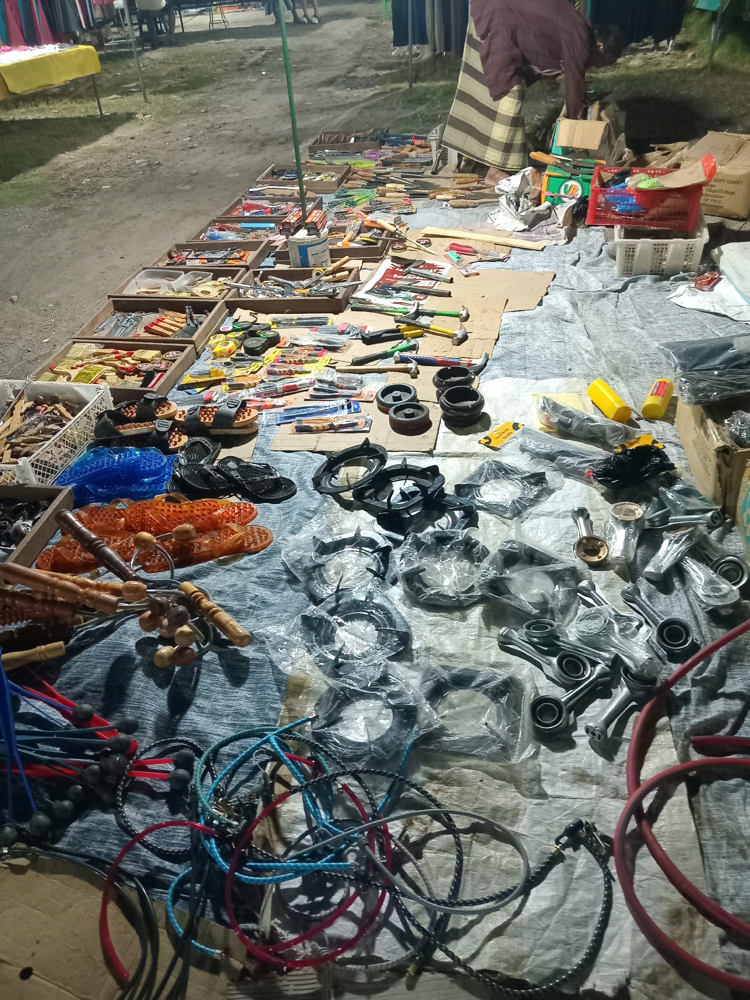 This is a nighttime, eye-level, full-shot photograph of a street market stall in Sedati Agung, Indonesia. The stall is set up on a dirt and gravel ground covered with a light-colored tarp. The foreground is dominated by various items displayed on the tarp, including: a collection of assorted tools such as hammers, pliers, and screwdrivers arranged in wooden trays; several pairs of sandals and flip-flops; a blue plastic mat; and a variety of metal objects, some of which are bagged in plastic, appearing to be stove parts or mechanical components. There are also some coiled ropes or cables in various colors. In the background, the market continues with more stalls, some covered with tarps and displaying goods, and the silhouettes of people are visible. The overall scene depicts a bustling nighttime market environment.
