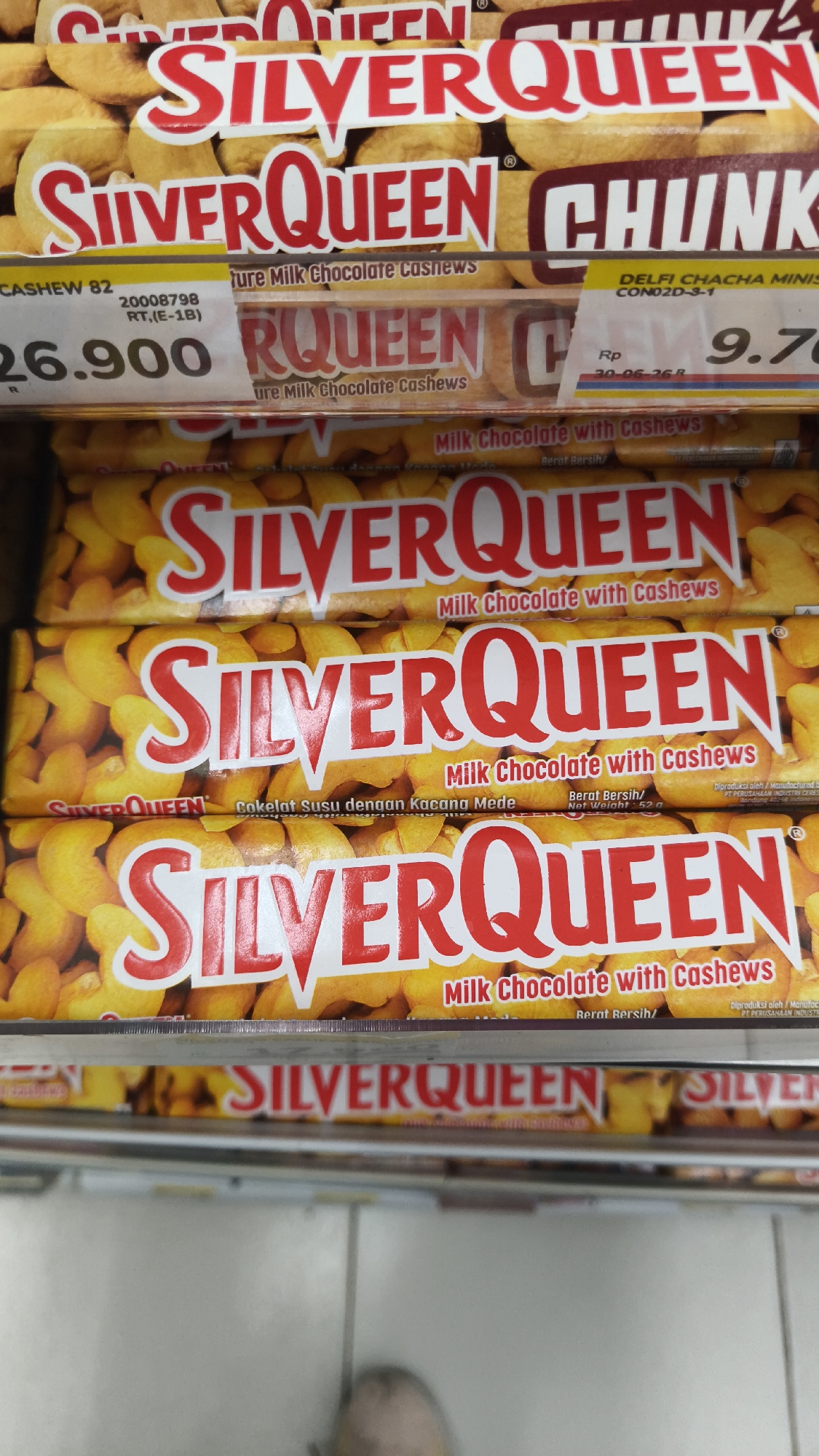 The image shows a retail display of SilverQueen chocolate bars, a popular brand in Indonesia. Several bars are stacked on shelves, with the branding and cashew nut imagery clearly visible. The top shelf displays larger bars labeled "SILVERQUEEN CHUNKS." Below that, two shelves feature smaller bars, one with text in Indonesian ("Cokelat Susu dengan Kacang Mede") and another with English text ("Milk Chocolate with Cashews"). Price tags are visible on some of the products. The overall environment appears to be a supermarket or convenience store in South Jakarta, Indonesia, given the product branding and typical retail setting.