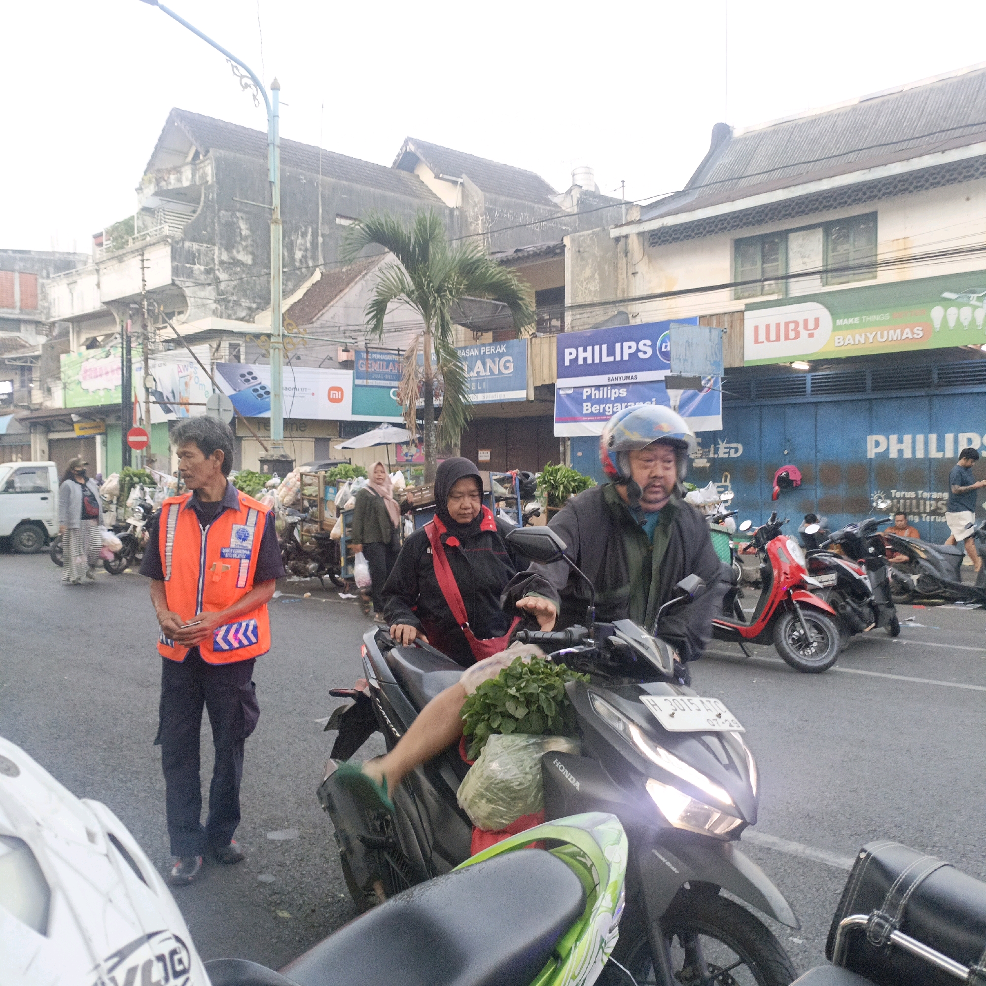 The image depicts a street scene in Salatiga, Indonesia, likely in a market or commercial area. The focal point is a man in the foreground wearing an orange vest with reflective stripes and dark pants, holding a smartphone. Behind him, a woman wearing a black hijab and jacket, with a red bag slung across her chest, is on a dark-colored motorcycle. She is holding onto the handlebars and appears to be stopped. A man in a helmet is also on a motorcycle behind her. In the background, various buildings with signs are visible, including "PHILIPS" and "LUBy". Other individuals, motorcycles, and market stalls with produce are also present, suggesting a busy urban environment. The overall lighting suggests it might be early morning or late afternoon.