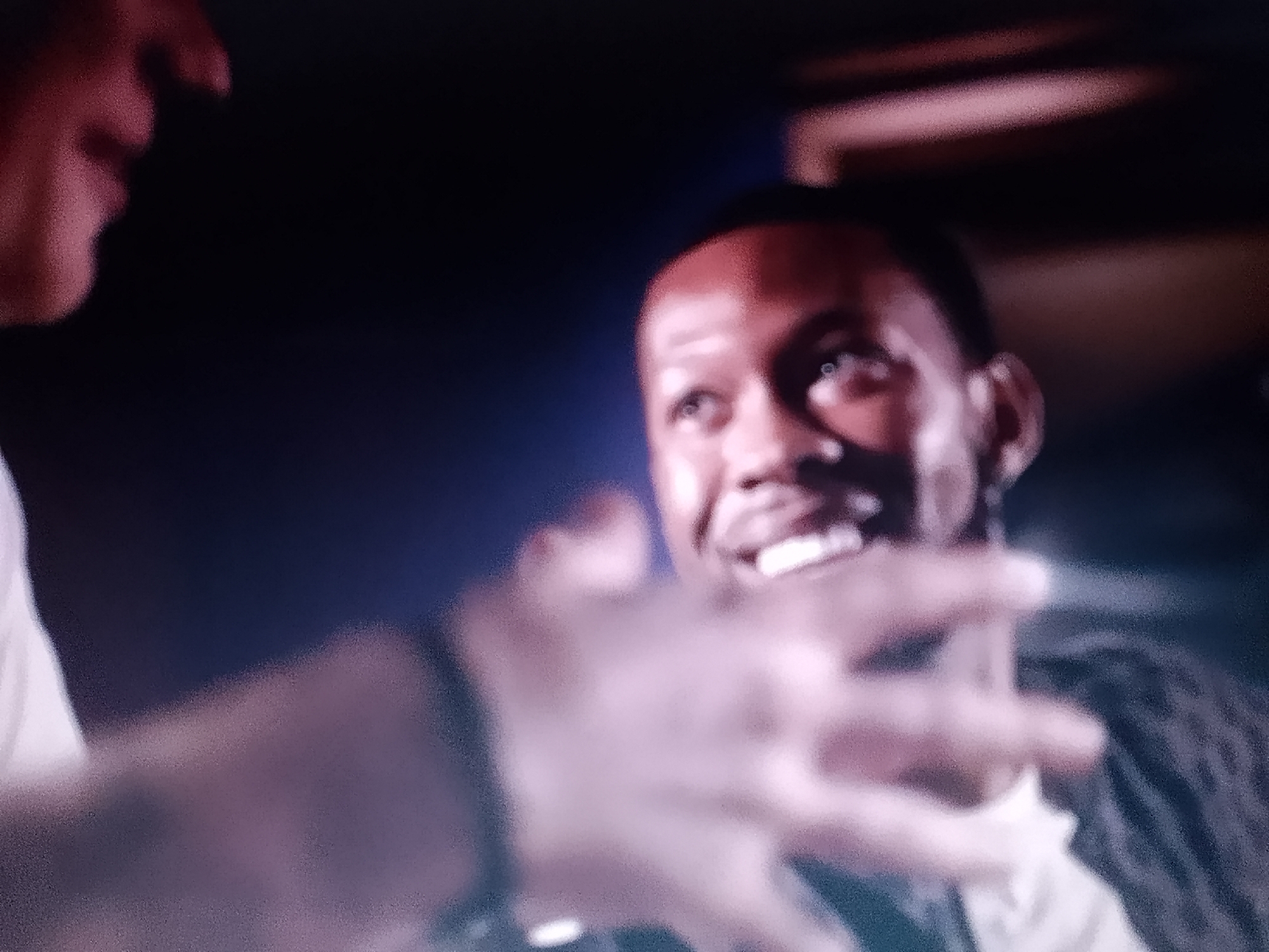 A medium indoor shot depicts two men. One is partially out of focus on the left, showing part of his head, left shoulder, white shirt, left hand, and left wrist adorned with a black wristband. The other man is more sharply in focus to the right. He has short dark hair, a light complexion, and is smiling, exposing white teeth. His expression is friendly, and he appears to be in a casual or conversational pose. He is wearing what appears to be a dark-colored jacket or shirt over a white undershirt. The background is dark, with a hint of reddish-brown above the man's head on the right. The setting and context provide no direct indicators of Adankolo, Nigeria.