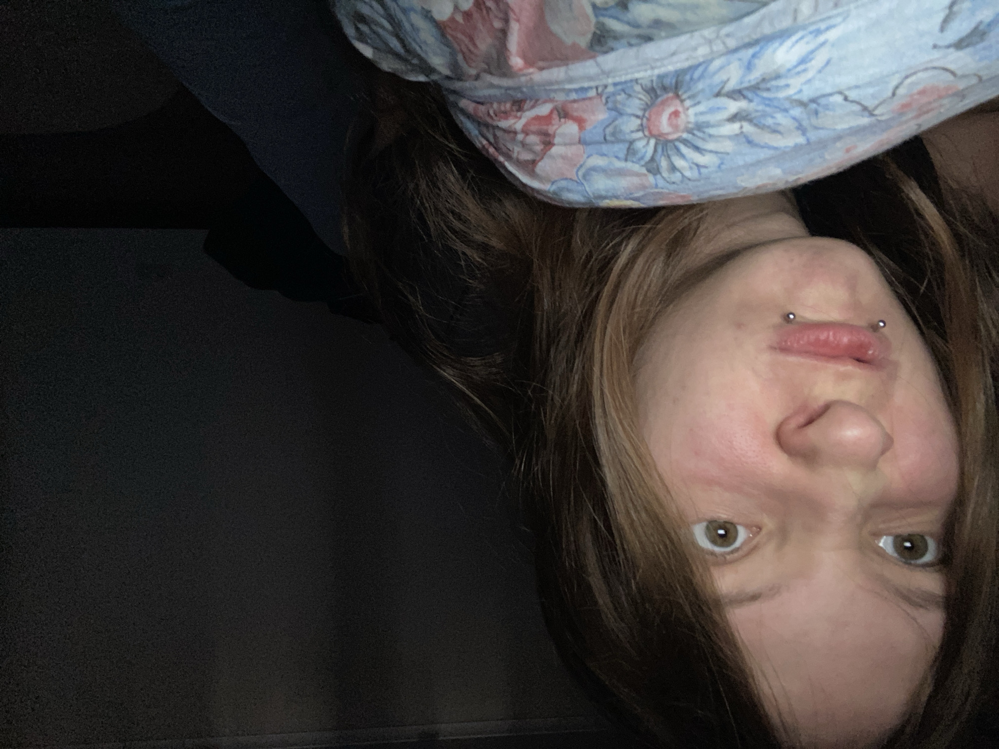 The image is a close-up, self-taken photograph of a young person with light brown hair, brown eyes, and visible lip piercings. The person is making a pouting or scrunched-up facial expression. They are partially covered by a blue, white, and pink floral patterned blanket or sheet. The background is dark and out of focus, with a faint hint of a black surface on the right. The lighting suggests an indoor environment, possibly a bedroom. There is no discernible indication of the specific location within Tolyatti, Russia.