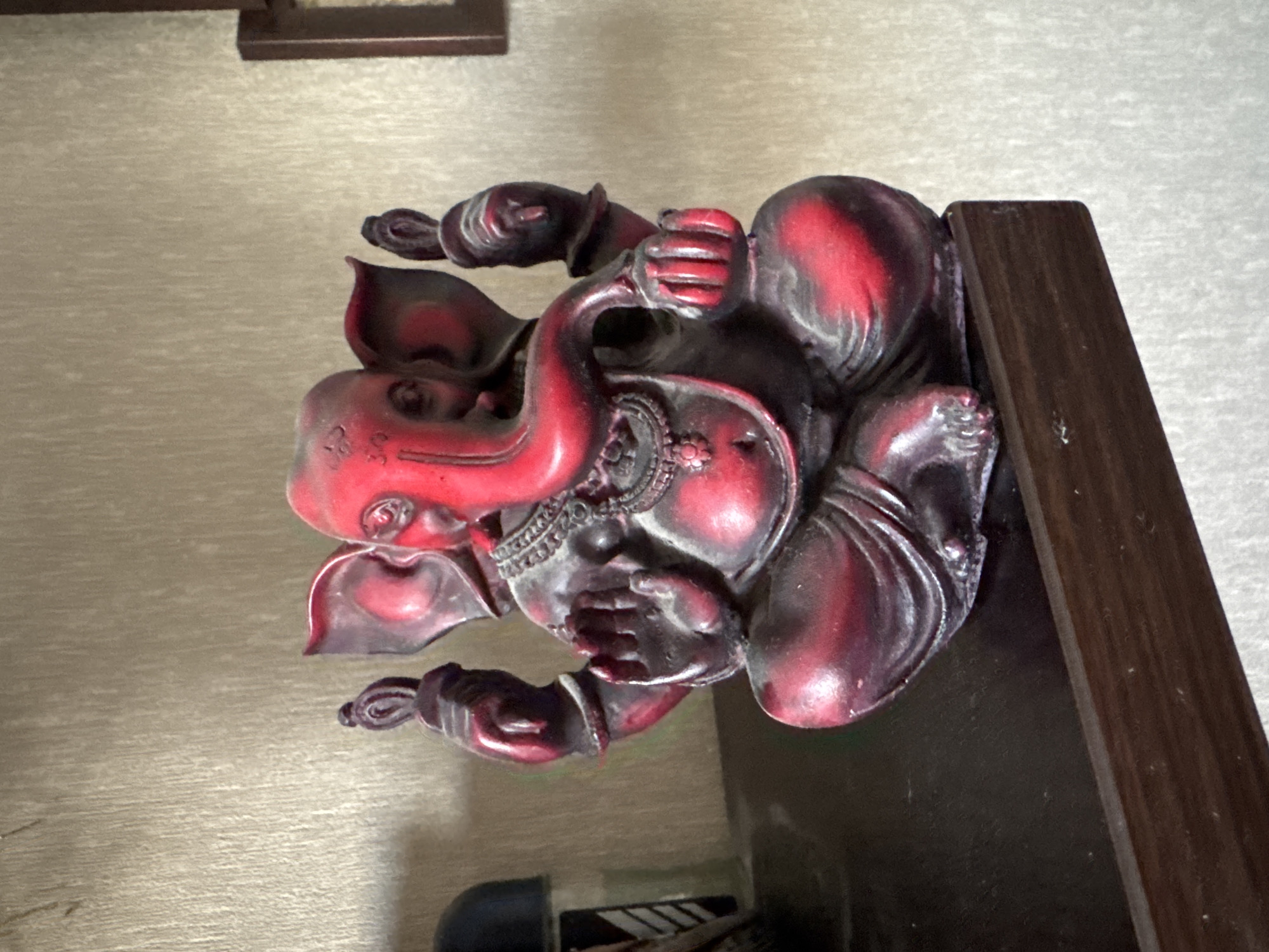 A red and black statue of Ganesh, the Hindu deity, is seated on a dark wooden shelf against a textured beige wall. The statue depicts Ganesh with his characteristic elephant head, four arms, and ornate jewelry. The Ganesh statue is the primary subject of the image. The environment appears to be an indoor setting, possibly a home or a shop, in Bishkek City, Kyrgyzstan. No people are present in the frame.