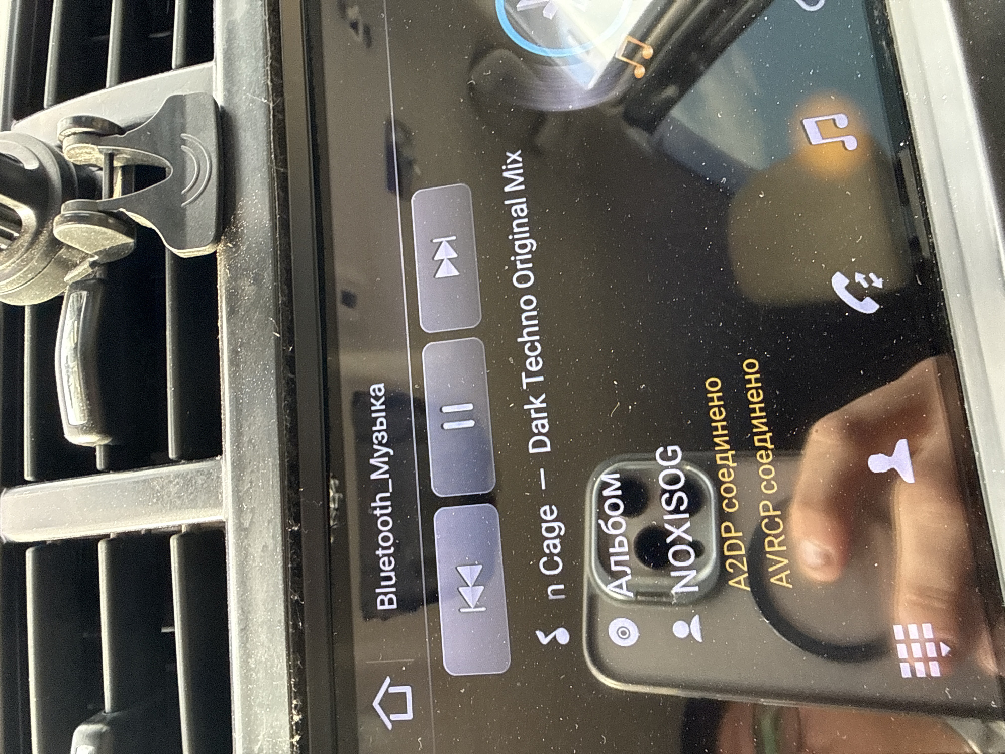 A vertical, close-up, eye-level shot shows a car's infotainment system screen mounted on a black car vent. The screen displays music playback controls, song title, and album information in Russian. The visible text includes "Bluetooth_Музыка," "Sn Cage -- Dark Techno Original Mix," "• Альбом," "NOXISOG," "A2DP соединено," and "AVRCP соединено."
On the left side of the screen, there are music playback buttons: a skip backward button with double arrows pointing left, a pause button with two vertical bars, and another button with a double arrow pointing right, possibly for skipping forward. A house icon is visible at the bottom left of the screen, indicating a home screen or main menu.
To the right, the album title "NOXISOG" is displayed below the word "• Альбом," which translates to "Album." The status of Bluetooth connections is shown as "A2DP соединено" and "AVRCP соединено," meaning "A2DP connected" and "AVRCP connected."
A finger is partially visible at the bottom right of the screen, suggesting user interaction. A silhouette of a person is present on the right side, possibly representing a user profile or contact icon. Another icon at the bottom right of the screen resembles a grid of squares, likely indicating a menu or navigation option.
The screen is illuminated and shows a bright reflection of something, possibly the interior of the car or a person's face. The screen and surrounding car vent are dusty, indicating the car may be in use or has been in storage. The overall scene depicts the user interface of a car's multimedia system during music playback via Bluetooth. The image lacks specific time of day or weather cues, but the context is clearly within a vehicle.