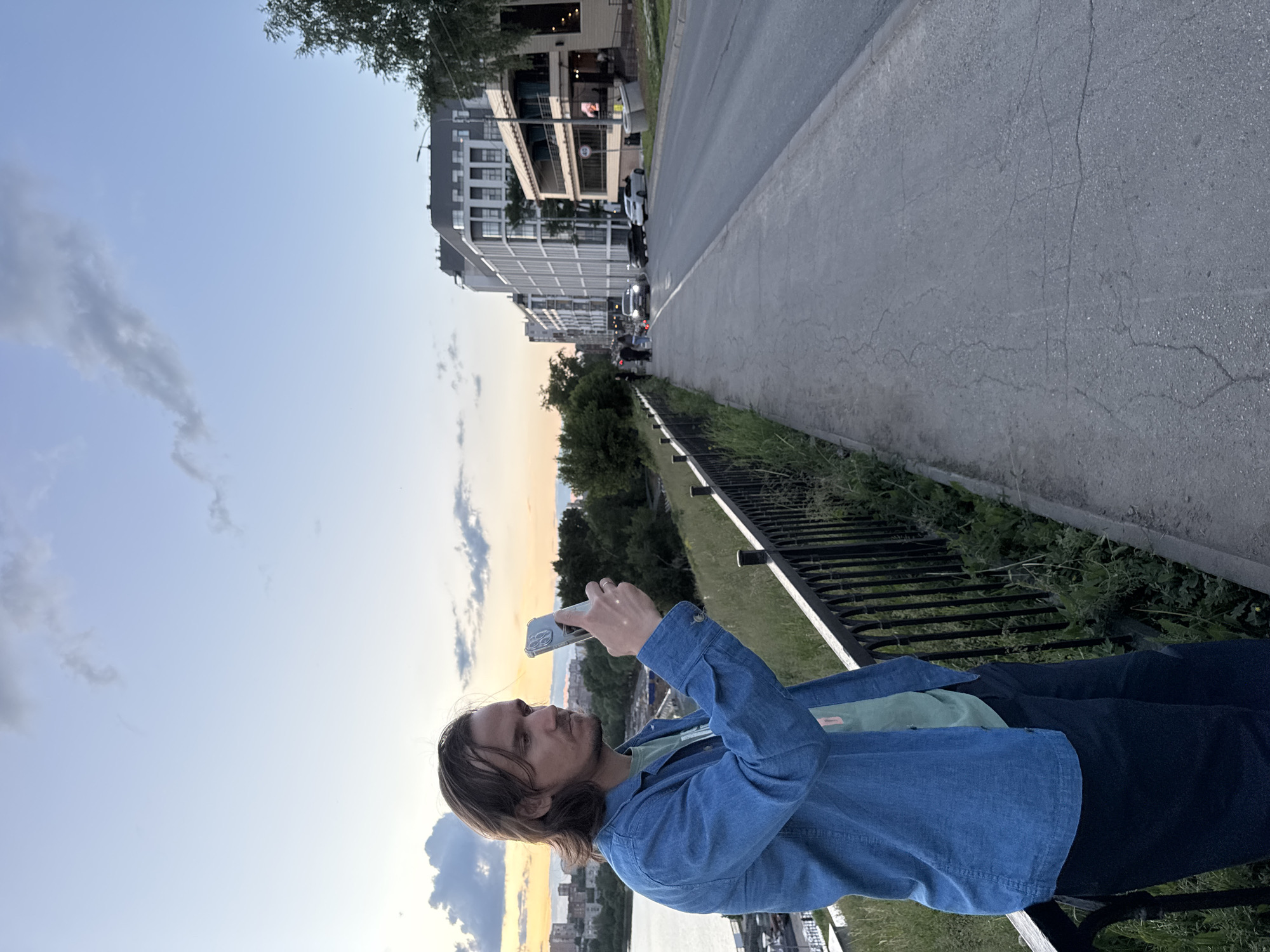 The image, captured in Izhevsk, Russia, features a man standing on an elevated path or promenade during what appears to be sunset or late evening. He is positioned on the left side of the frame, facing right, holding a smartphone with a pop socket in both hands, suggesting he is taking a picture or video of the scenic view. He wears a blue denim jacket over a light green t-shirt and dark pants, with medium-length brown hair.
A black metal railing runs along the edge of the promenade, separating the man from the scene below. To the right, an asphalt street, showing some cracks, stretches into the distance, bordered by a strip of grass and scattered vegetation. Modern multi-story buildings with various architectural details, including balconies and prominent windows, line the far side of the street. Several cars are parked or driving on the street, and a few distant pedestrians are visible. Beyond the urban structures, a wide body of water, possibly a river or lake, is visible under a sky painted with the warm, soft colors of twilight, transitioning from light blue overhead to shades of orange and yellow near the horizon. Sparse clouds are scattered across the sky, contributing to a serene atmosphere. No legible text is visible in the frame.