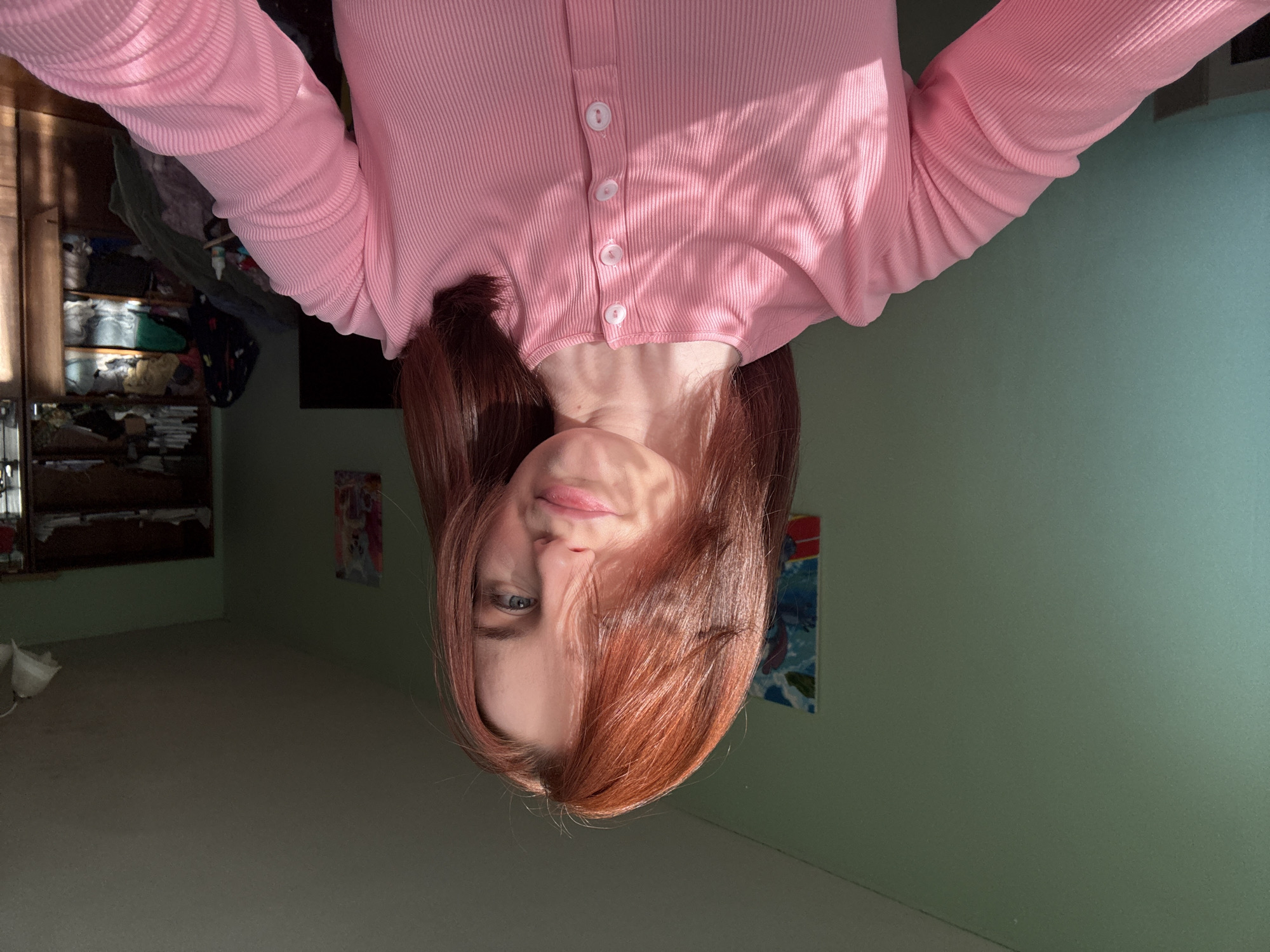 A reddish-haired woman wearing a pink, button-up shirt is upside down in a room with light green walls. Two framed pieces of art hang on the wall, with a wooden cabinet visible in the distance to the left. The foreground is a carpeted floor. Location is unknown.