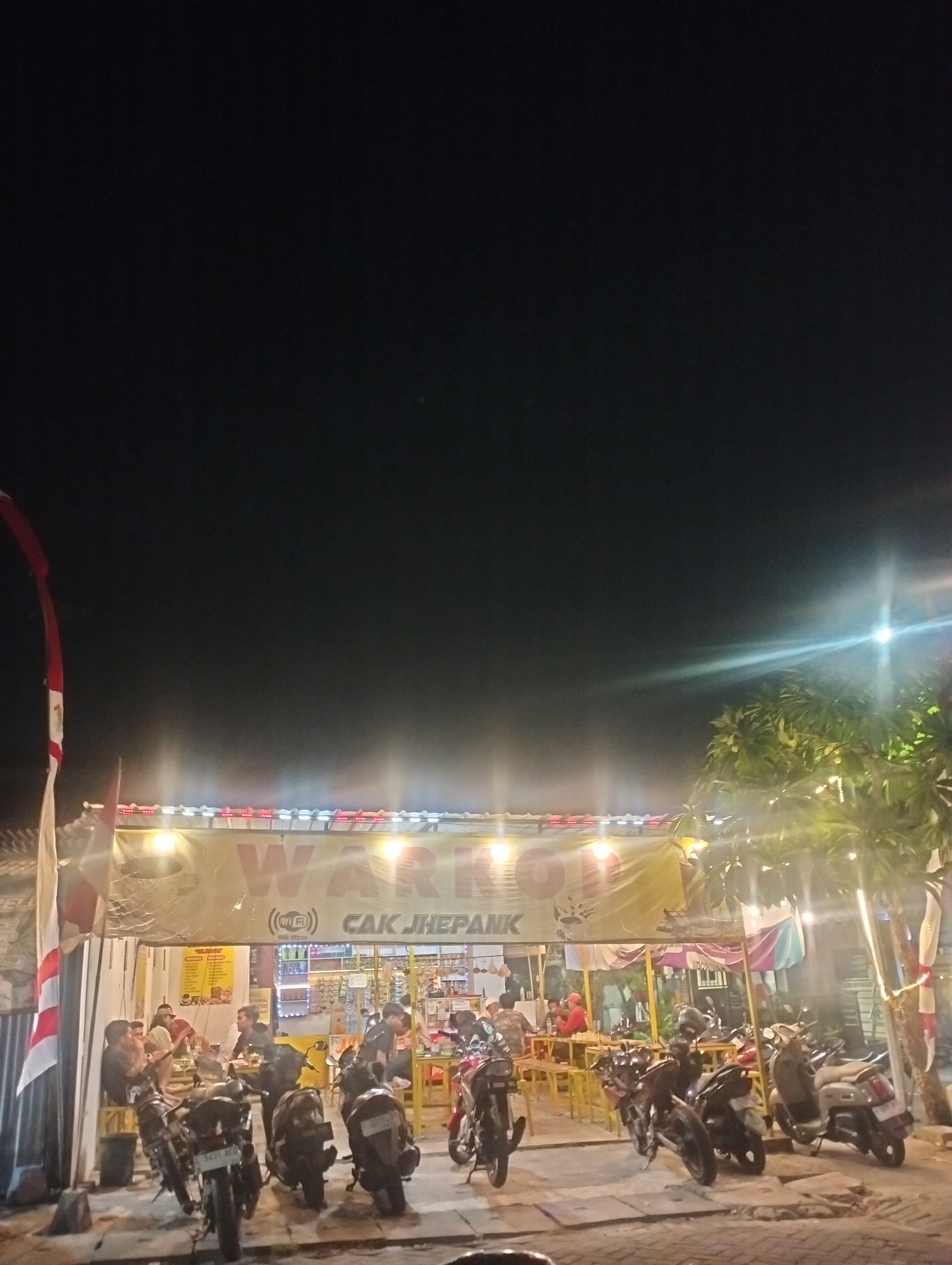 The image captures a street-level view of a warung (small shop/eatery) at night in Surabaya, Indonesia. The warung has a large yellow banner overhead with red lettering that appears to say "WARUNG CAK JHEPA". Below the banner, there are lights illuminating the interior of the warung. Inside, several people are visible sitting at tables, suggesting a dining or social gathering.
In front of the warung, numerous motorcycles are parked. The street in front of the warung is paved, and there are lights strung along the warung's exterior, creating a warm glow. A tree with decorative lights is visible to the right of the warung. A flag, partially visible on the left, has red and white stripes. The overall impression is that of a lively local establishment at night.