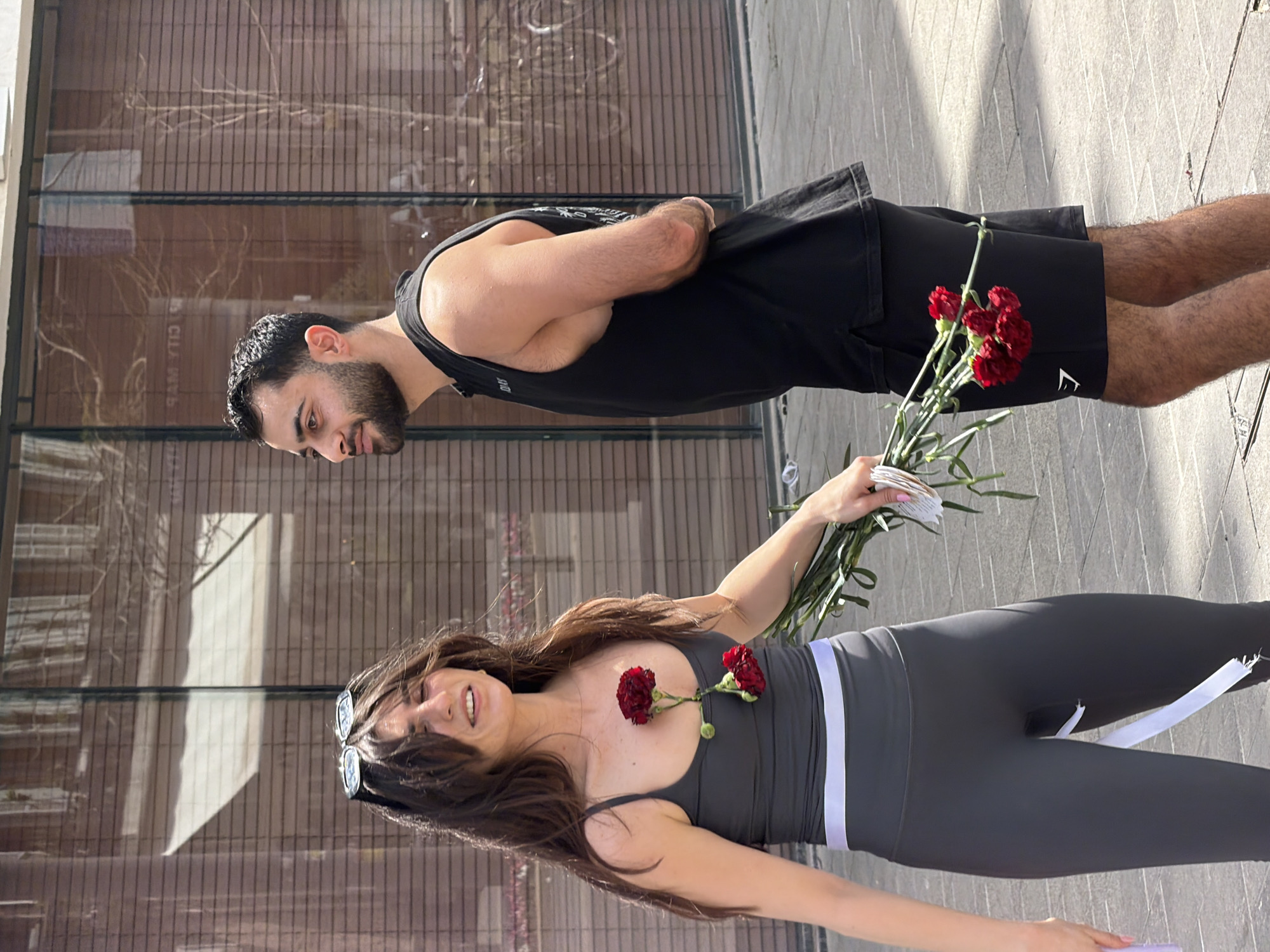 An outdoor, eye-level shot captures a man and a woman on a sunny day in an urban environment, identified as Tel-Aviv, Israel. The woman is in the foreground, smiling broadly and looking towards the camera. She has long dark hair and is wearing a dark gray or black sleeveless athletic top with a white stripe at the waist, and matching leggings. She holds a bouquet of several red carnations and a stack of small white cards or notes in her left hand. One red carnation is tucked into the neckline of her top.
Standing slightly behind and to the right of her is a man with dark hair and a beard, wearing a black sleeveless top and black shorts. His arms are crossed behind his back, and he is looking at the woman with a gentle expression. A white logo, featuring the letter "E" and a shark fin-like symbol (suggesting the brand Gymshark), is visible on his shorts.
The background consists of a modern building with large glass windows and horizontal dark panels. Reflections of bare tree branches and faint, reversed text "YTC RAM" are visible in the glass. The ground is paved with rectangular blocks, and strong shadows indicate bright sunshine, suggesting it is daytime. The scene conveys a happy, possibly romantic, moment or gift exchange.