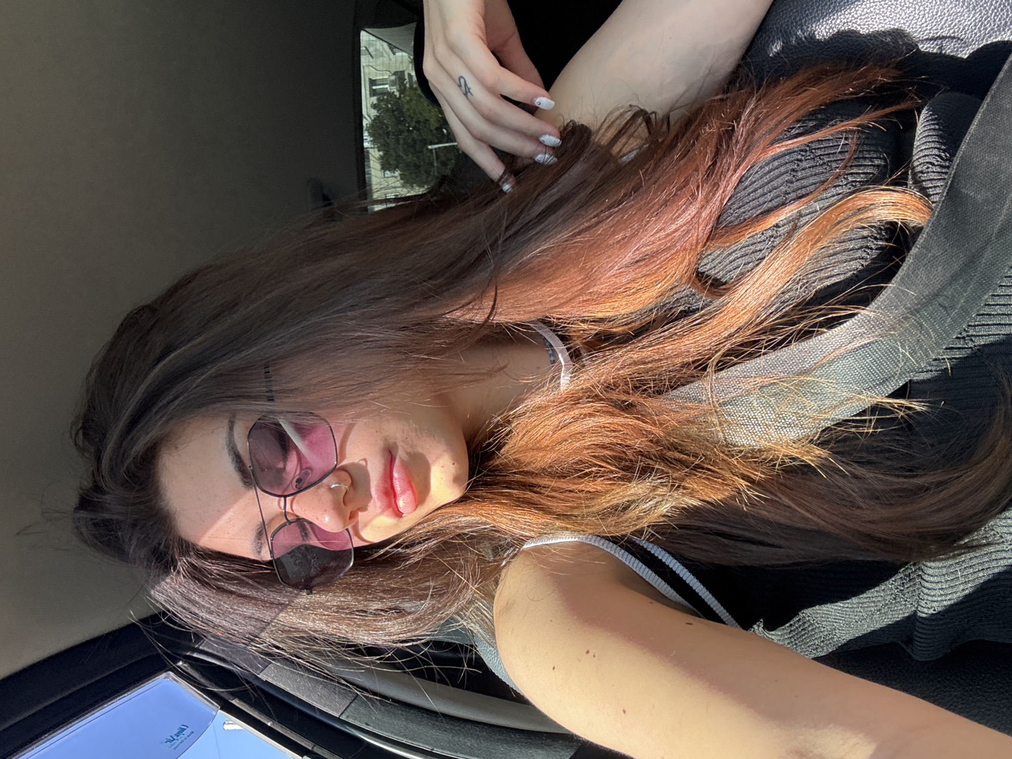 A young woman with long, dark brown hair is captured in a selfie taken inside a car. She is wearing large, hexagonal sunglasses with pink lenses and a black ribbed top with white trim. Sunlight is streaming in from the left side of the frame, casting a warm glow on her face and hair, and creating shadows on her arm and clothing. A seatbelt is visible across her chest. Her right hand is resting on her shoulder, revealing manicured fingernails and a small tattoo on her finger. The background outside the car window shows a building and some trees, suggesting an urban setting, possibly Sofia, Bulgaria, during daylight.