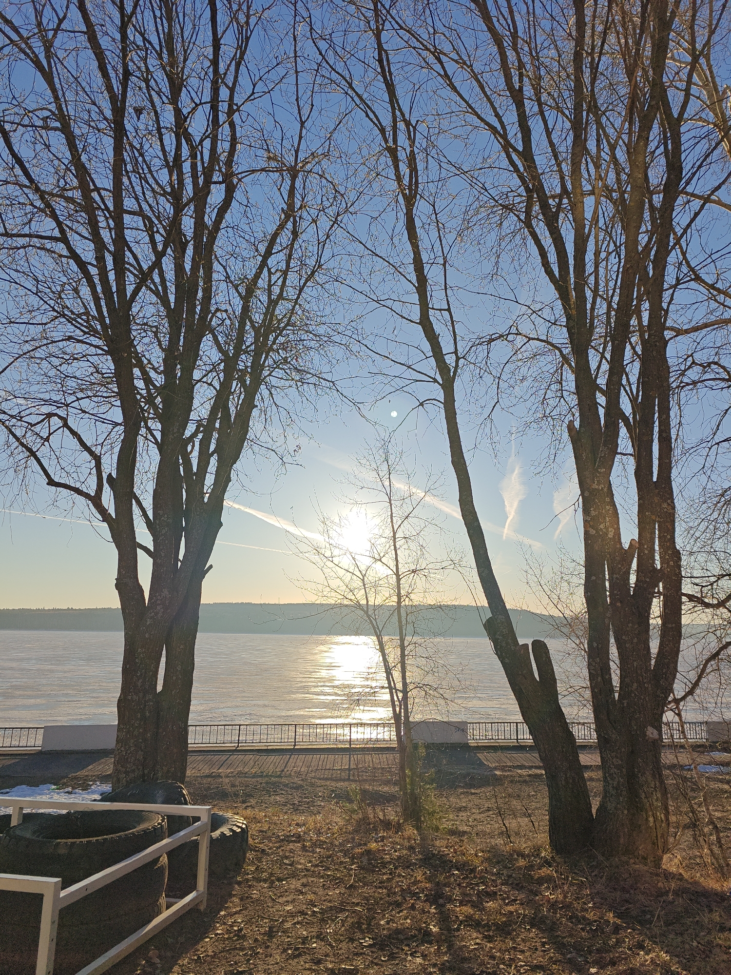 The image shows a waterfront view in Chaikovsky, Russia, at what appears to be sunset or sunrise. A body of water, potentially a lake or river, is partially frozen or covered with ice, reflecting sunlight. A metal fence runs along the waterfront promenade. Trees with bare branches frame the view. In the foreground are tires within a white painted frame, possibly part of a playground or park feature. The ground is covered with dry grass and leaves. Contrails are visible in the sky.