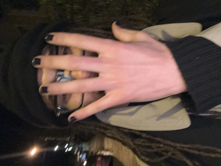 A person covers their face with a hand, revealing only their eyes and glasses. The hand has dark nail polish. The person wears a black hooded garment and a lighter-colored jacket or shirt beneath. Streetlights and blurred buildings are visible in the background, suggesting an urban environment at night. The image is slightly tilted, adding a sense of unease.