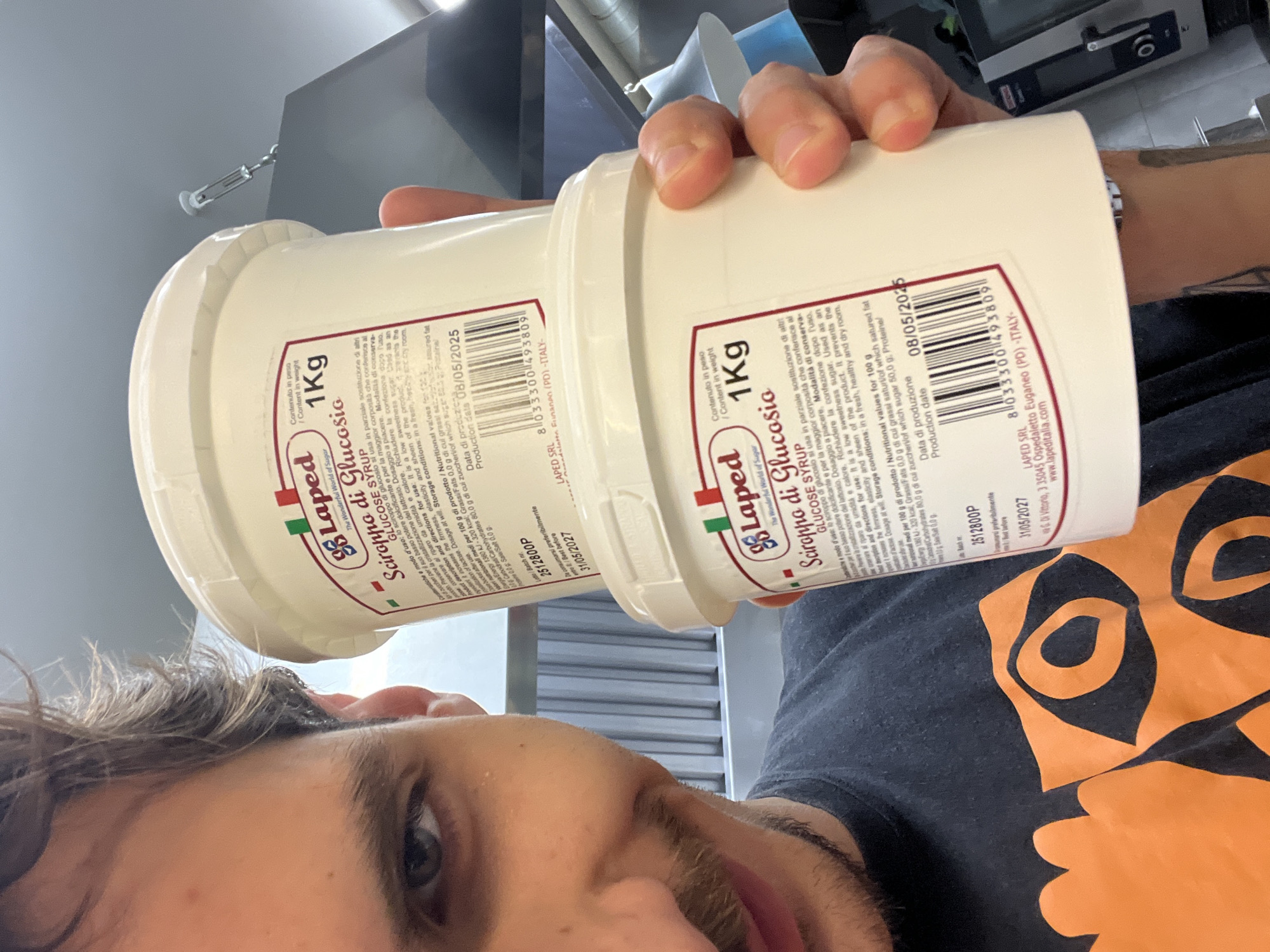 The image shows a man holding two 1 kg tubs of "Sciroppo di Glucosio" (Glucose Syrup) produced by "Laped SRL" in Italy. The tubs have production dates of 08/05/2025 and a best-before date of 31/05/2027. The man is visible from the chest up, wearing a black t-shirt with an orange design. He is looking towards the camera. The background appears to be a commercial kitchen setting with stainless steel surfaces and equipment. The location is not specified as Budva, Montenegro in the image.