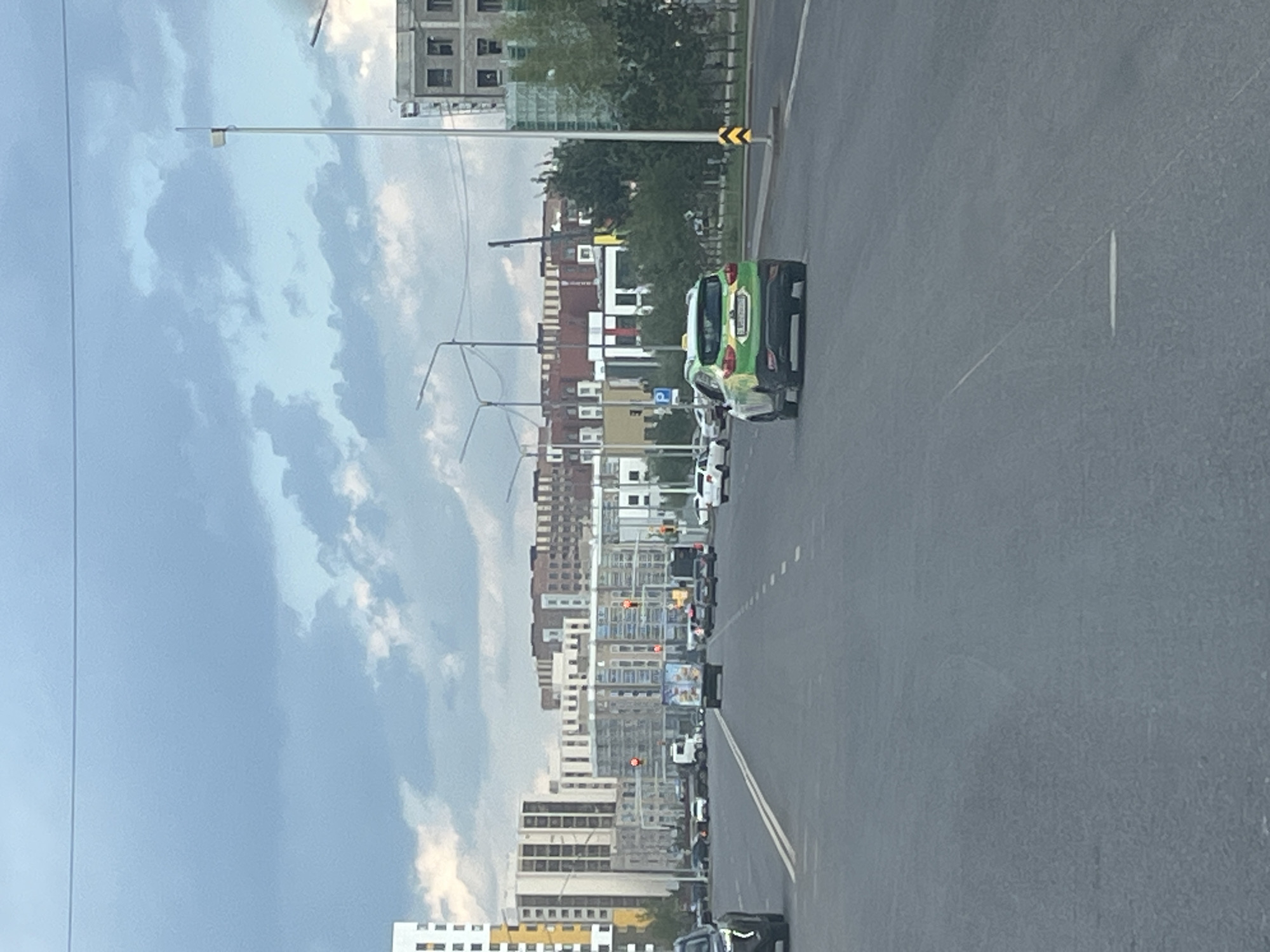 The image depicts a street scene in Astana, Kazakhstan, under a cloudy sky. The foreground is dominated by a grey asphalt road with white lane markings. Several vehicles are visible on the road, including a green and yellow taxi in the right lane, a black SUV partially visible on the left, and various other cars and trucks further down the road. In the background, modern buildings of varying heights and architectural styles line the street, some featuring glass facades and balconies. There are also streetlights and traffic signals visible. Trees and some construction scaffolding add to the urban environment. The overall impression is of a busy city street during daytime, likely with overcast weather.