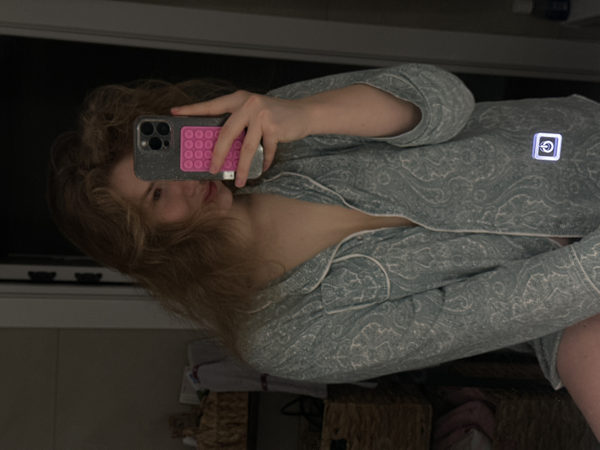 A woman is taking a selfie in a mirror. She is wearing patterned pajamas. Her phone is a dark color with a pink PopSockets grip. The reflection shows part of a room with a black object above her, likely a television. There are baskets beneath the mirrored surface, possibly in a bathroom or bedroom. There is no information to suggest the location is in Kazan, Russia.