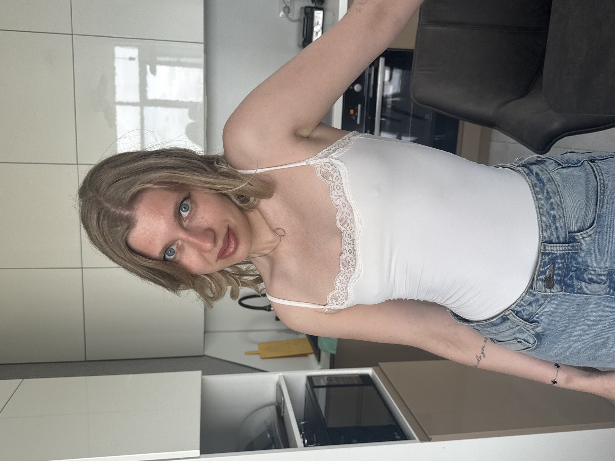 A young woman with blonde hair and blue eyes is standing in a modern kitchen. She is wearing a white tank top with lace trim and denim overalls. The kitchen has white cabinets and a dark gray chair is partially visible in the background. A microwave and other kitchen items are visible in an open drawer. There is no information to verify the location in the city of Rostov-on-Don, Russia.