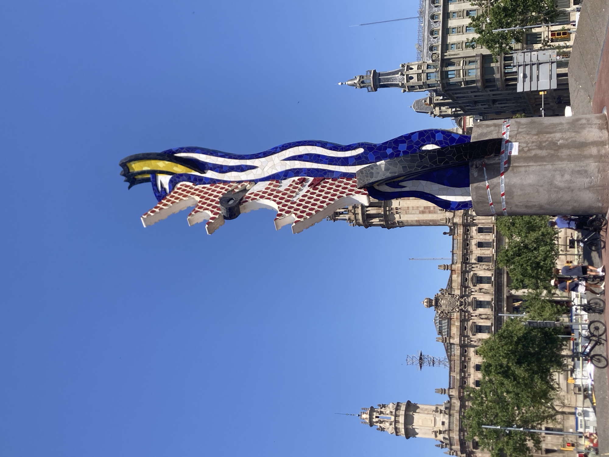 This is a full-shot, eye-level, outdoor photograph taken on a bright, sunny day with a clear blue sky. The primary subject is a tall, abstract sculpture made of brightly colored mosaic tiles, predominantly blue, white, yellow, and red dots. The sculpture is positioned in the foreground on the right side of the frame, its base made of rough concrete and wrapped with red and white caution tape.
In the background, several large, ornate buildings with distinct architectural styles are visible. The building on the left features a prominent tower with intricate detailing. The central building is grand, with a symmetrical facade and a large coat of arms above its entrance. The building on the right has a darker facade with a stepped roofline.
A few people are present, walking and standing near bicycles in the middle ground, to the left of the sculpture. Green trees are scattered throughout the scene, lining the street and surrounding the buildings. The overall environment suggests an urban setting, likely a public plaza or square in a city. Based on the sculpture, "Cranes" by Claes Oldenburg and Coosje van Bruggen, and the surrounding architecture, the location is likely the Port Vell area of Barcelona, Spain, near the Columbus Monument and the waterfront.