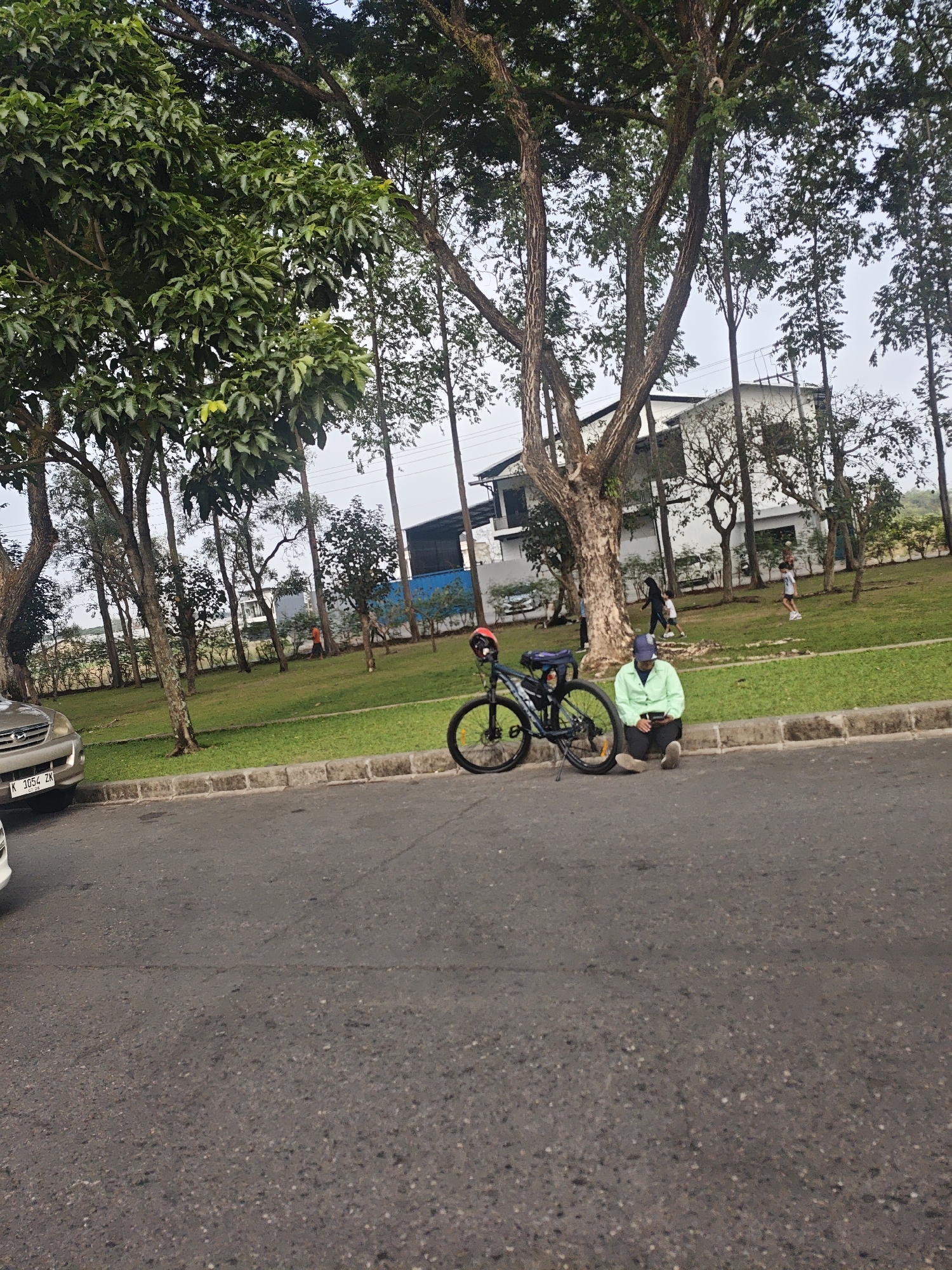 The image depicts a scene on a paved road in Bacin, Indonesia, with a grassy area and trees to the right, and buildings in the background. In the foreground, a bicycle is parked next to a person sitting on the curb. The person is wearing a light green long-sleeved shirt and dark pants, with a blue cap. They are holding a smartphone and looking down at it. The bicycle is dark colored with a blue bag attached to the back. A red helmet is resting on the handlebars. To the left of the bicycle, a silver car is parked on the side of the road. The car has a license plate with the characters "K 1054 ZK". The grassy area features several trees, and in the distance, a white building with a dark roof is visible. There are other individuals walking in the background, including a person in an orange shirt and two children. The overall lighting suggests it is daytime.
