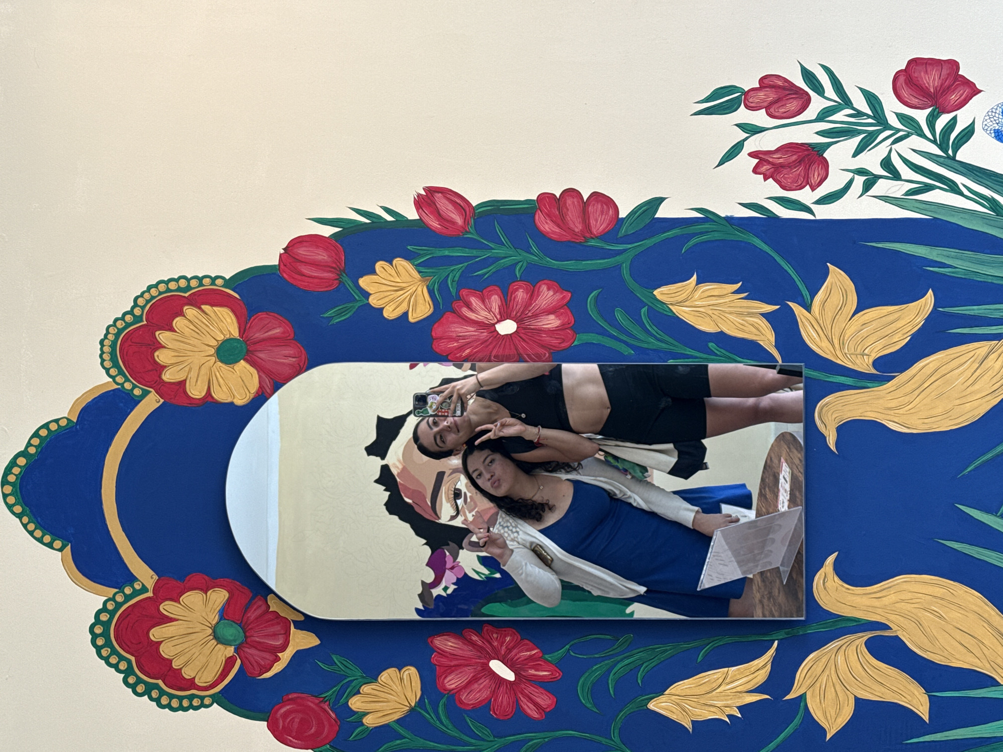 Two young women are reflected in a mirror positioned against a painted wall. The wall features a large blue field adorned with stylized red, yellow, and green floral motifs. The mirror's frame is rectangular with rounded corners.
The reflection shows the women taking a selfie. One woman is wearing a black top and shorts, while the other is wearing a blue dress and a light-colored jacket. They are both making peace signs with their fingers.
The location is suggested to be Puebla, Mexico, based on the style of the artwork on the wall.