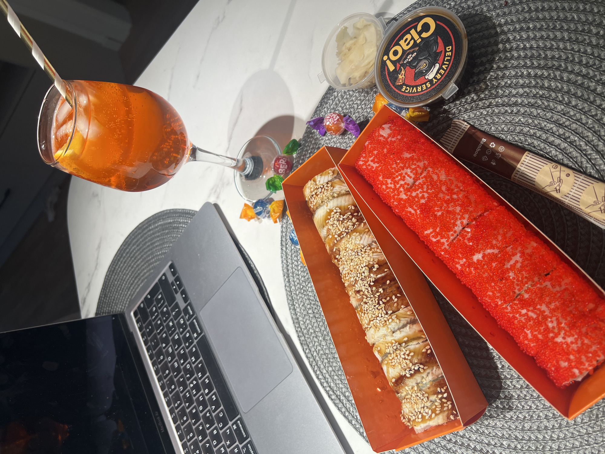 This media file captures an overhead view of a casual indoor setting in Budva, Montenegro, featuring food, drink, and a laptop. The scene is set on a white marble-patterned table, partially covered by a dark grey, woven placemat.
In the upper left, a wine glass filled with an orange-colored beverage, likely an Aperol Spritz, is visible. It contains ice cubes and a lemon or orange slice, with a white and striped paper straw inserted. Next to the glass, several small, brightly colored wrapped candies are scattered, one clearly showing "BER" on its wrapper.
Towards the right, two trays of sushi rolls are prominently displayed in orange cardboard containers. The left tray holds rolls topped with eel and sprinkled with sesame seeds, while the right tray features rolls generously coated with vibrant red fish roe (tobiko). Adjacent to the sushi, a clear plastic container holds pickled ginger, and another round black container with a yellow label reading "Ciao! DELIVERY SERVICE" and featuring a cartoon wolf is present. A brown paper packet, likely containing chopsticks, is visible to the right of the sushi, with the word "Bamboo" printed on it.
A silver MacBook Air laptop is open and positioned on the left side of the frame, showing part of its keyboard with both Cyrillic and Latin characters. The laptop's screen is dark and reflects some light.
The artificial lighting suggests it could be evening or simply an indoor scene with overhead lights, casting some soft shadows. No people are visible in the image. The arrangement suggests a relaxed meal or snack while using the computer, perhaps for work or entertainment.