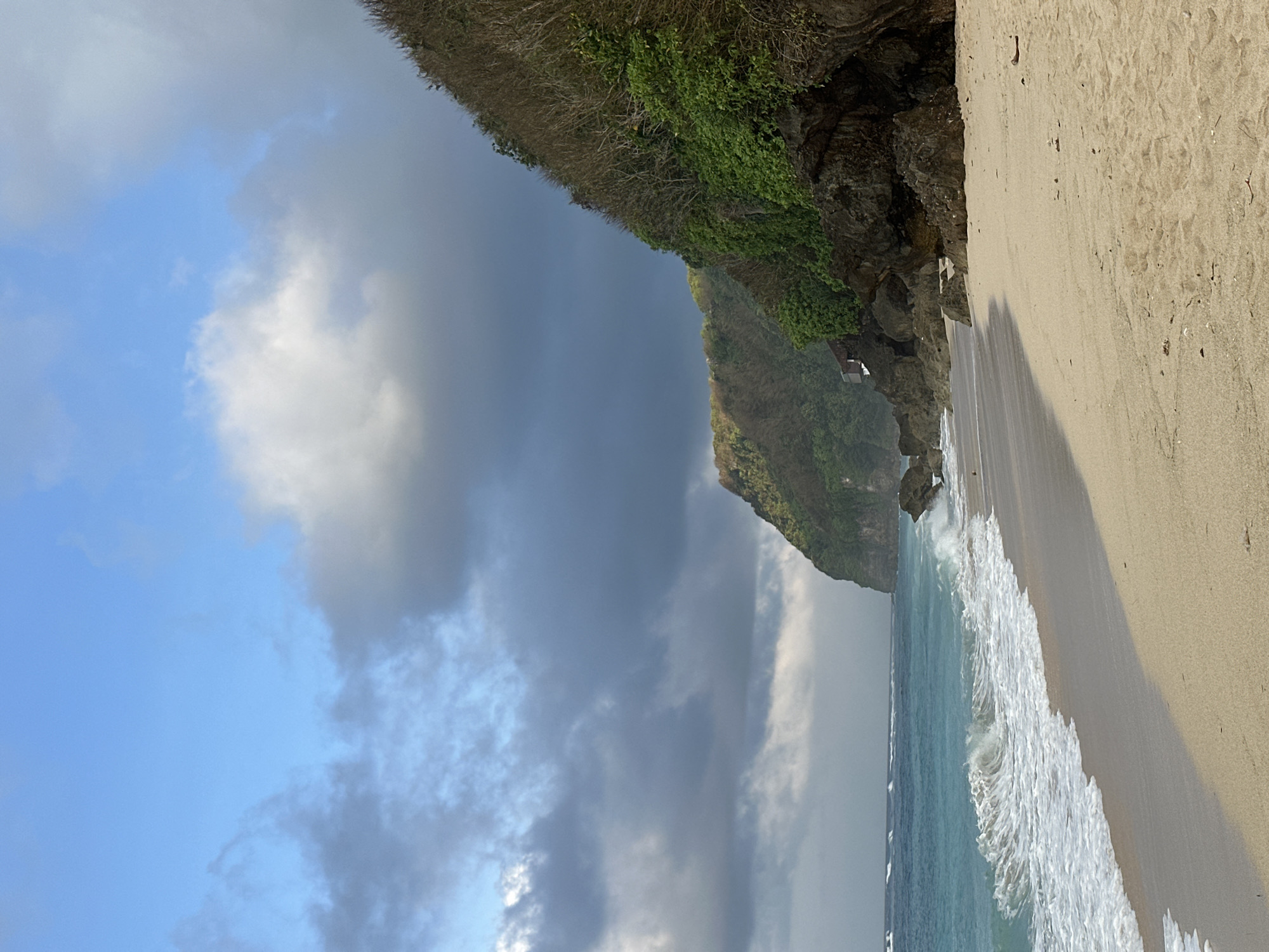 The image shows a sandy beach with turquoise water and white waves crashing on the shore. A lush green cliff rises from the sea on the right side of the frame, with some dry, bare branches visible on its upper slope. The sky is a mix of bright blue with scattered dark clouds. The sand is light brown and shows some footprints and scattered debris. A small, dark-colored structure, possibly a building or a hut, is partially visible nestled into the cliff face. The scene is devoid of any people or animals. The overall impression is a dramatic and somewhat moody coastal landscape. This scene is characteristic of many beaches in the Kuta Selatan region of Bali, Indonesia, known for its stunning cliffs and powerful surf.