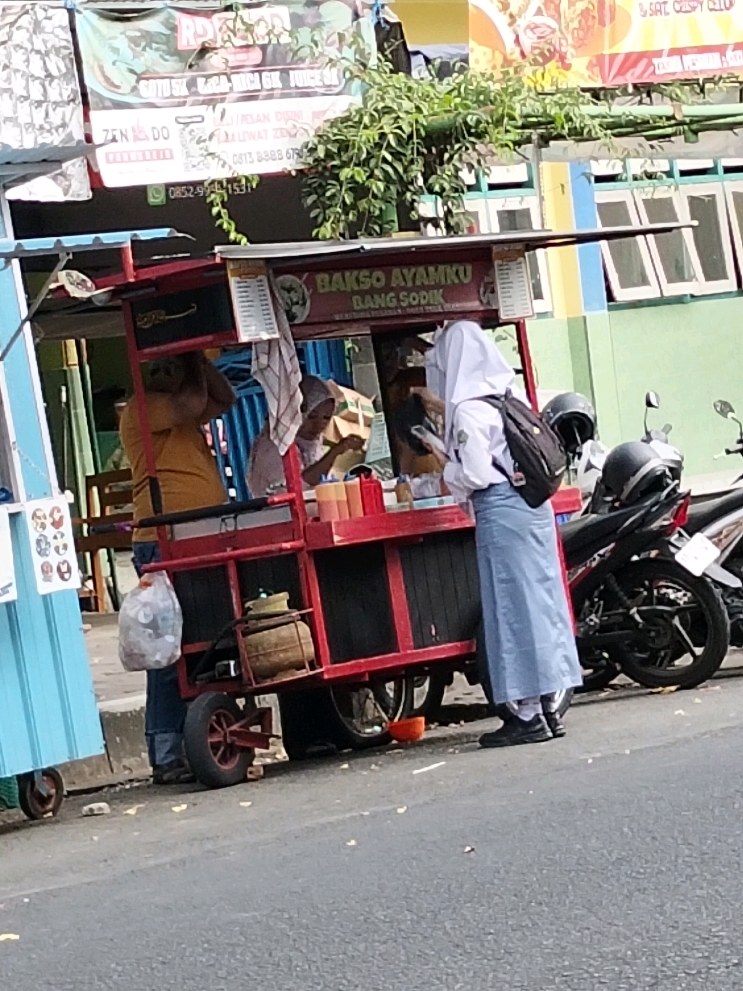 In Purworejo, Indonesia, a food cart with a red frame and black shelves is situated on the roadside. The cart displays a sign that reads "BAKSO AYAMKU BANG SODIK." A person wearing an orange shirt and blue pants stands behind the cart, holding a plastic bag. Another person, dressed in a white hijab, white shirt, and a light blue skirt, is interacting with the cart. They have a black backpack on their back and are holding a smartphone. Several motorcycles are parked behind the cart, some with helmets on them. The environment suggests an outdoor setting, likely a street, with greenery visible in the background. The ground is paved with asphalt.