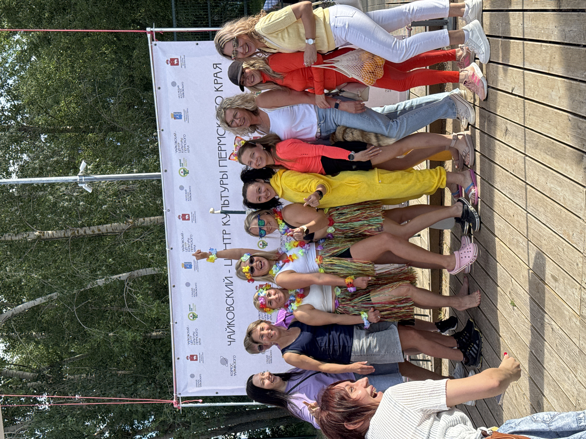 This media file shows a group of women posing for a photo in an outdoor setting, likely a festival or event. The women are standing in a line, facing the camera, and appear to be in good spirits, smiling and laughing. They are dressed in casual summer attire, with some wearing colorful outfits and flower leis, suggesting a festive atmosphere. One woman in the back is wearing white pants and a yellow top, while others are in orange, white, and yellow tops, paired with jeans and shorts. Some are also wearing Hawaiian-style skirts.
Behind the group is a large white banner with Cyrillic text and logos, which appears to be a promotional banner for the "Chaikovsky Center of Culture of Perm Krai." The banner is hung horizontally and appears to be part of the event's setup.
The ground beneath them is made of wooden planks, characteristic of a boardwalk or an outdoor stage. Sunlight suggests it is daytime, likely a warm and sunny day. The background shows trees and foliage, indicating an outdoor location. The overall impression is one of a joyful and celebratory gathering.