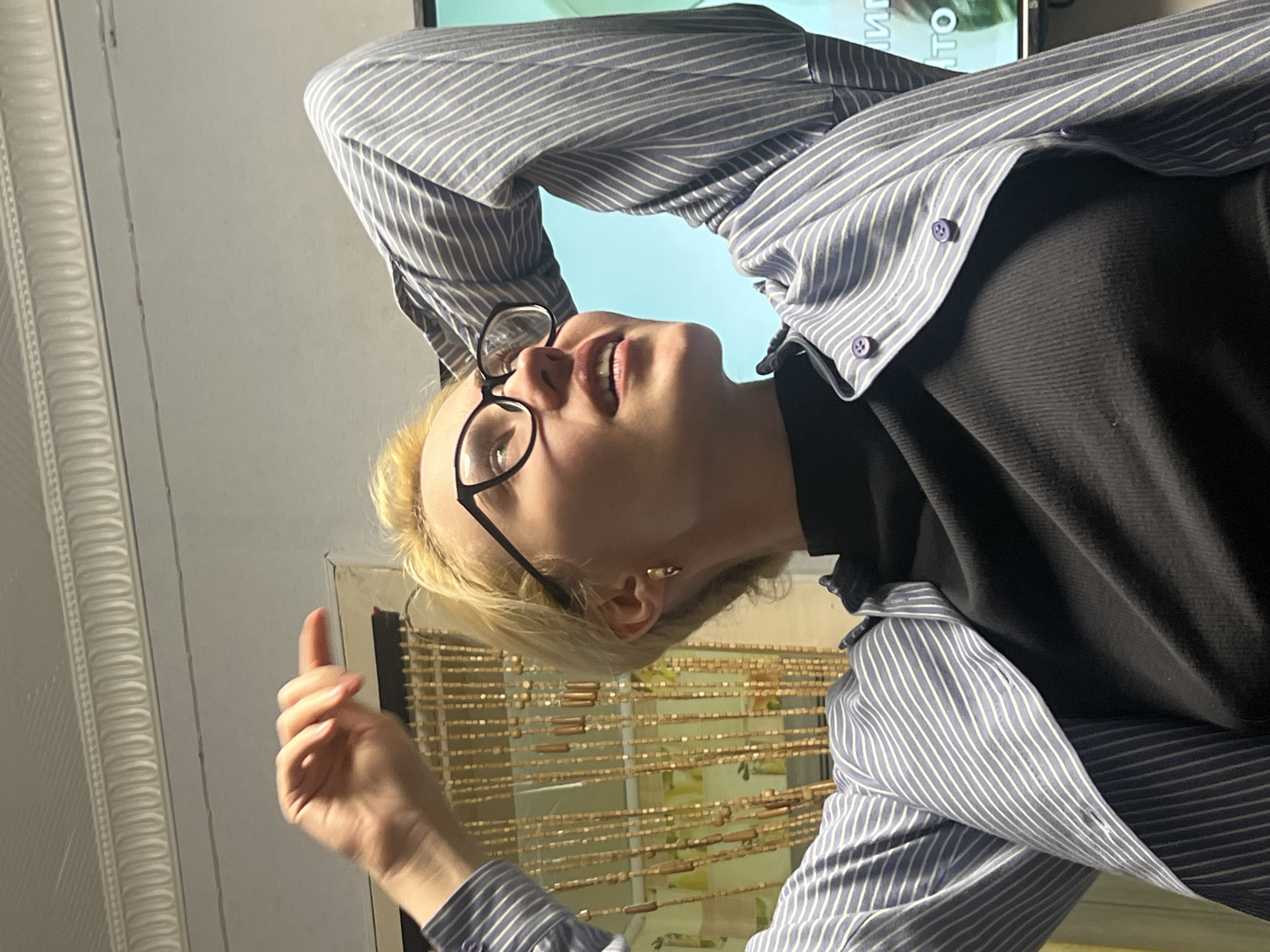 A person wearing glasses and a striped shirt stands indoors. They have blond hair and are wearing a black shirt underneath the striped one. Their left hand is raised. A beaded curtain hangs behind them. The location is Chaikovsky, Russia.