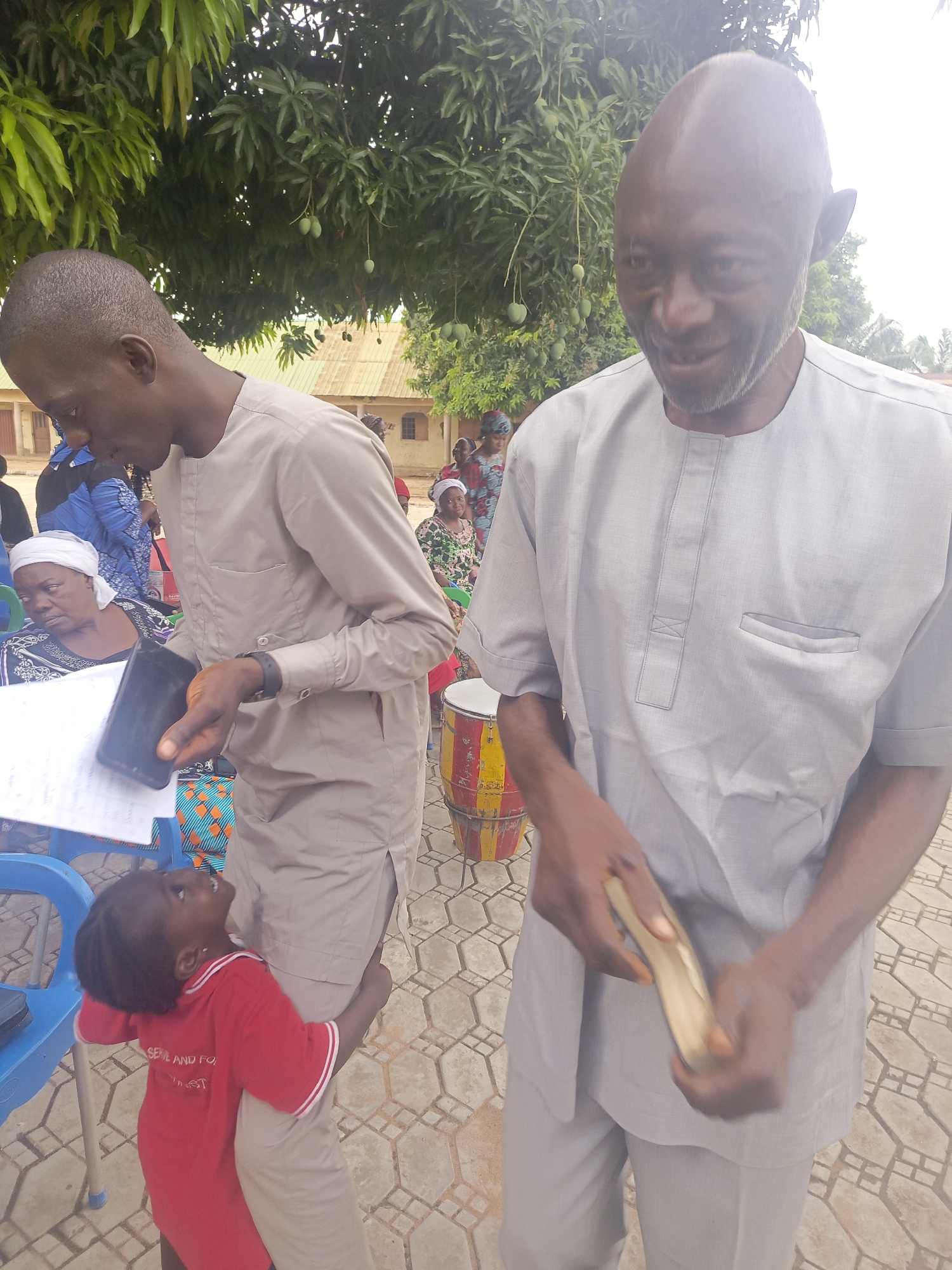 Akwanga, Nigeria: Two adult men, presumably related, stand on a geometrically-patterned patio. One man, balding and older, is holding a wooden object. The other, younger, is glancing at a phone or small tablet. A child wearing a red shirt clings to the younger man. In the background, other adults are seated and a drum is visible. Trees overhang the scene. Buildings are also visible in the background.
