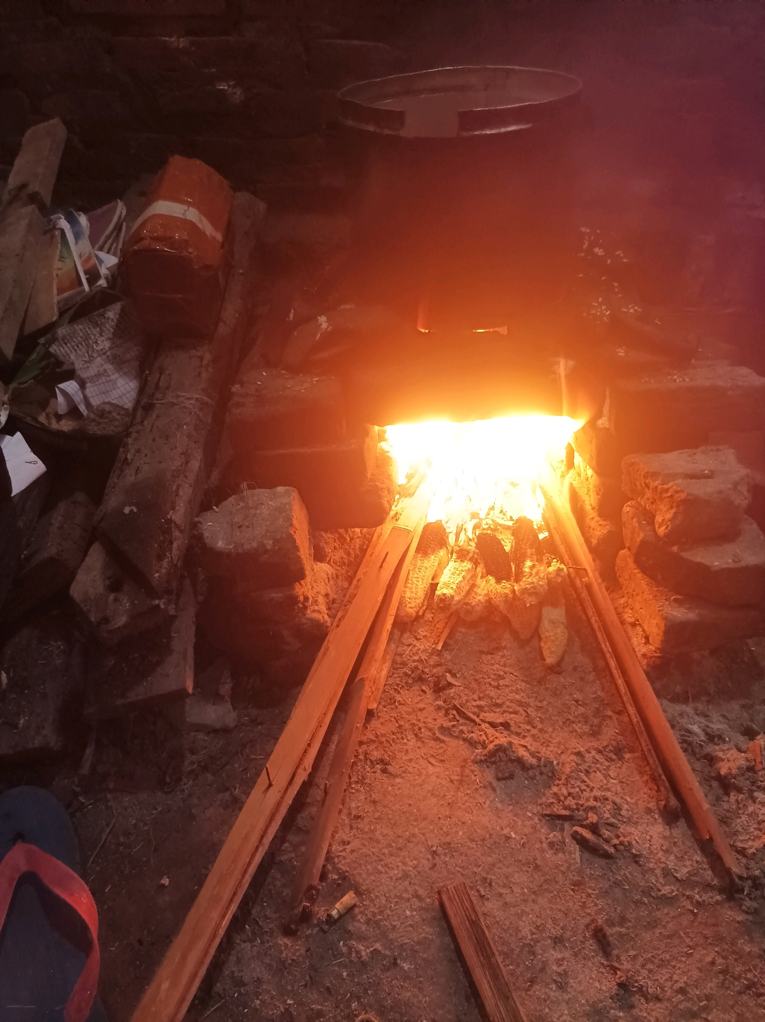 The image shows an outdoor, dimly lit scene with a fire pit in a rural or semi-rural setting, likely in Canga'an, Indonesia. The fire is burning brightly within a brick structure that appears to be an outdoor stove or hearth. Several pieces of corn on the cob are positioned over the fire, suggesting they are being roasted. Around the fire pit, there are various objects, including bricks, pieces of wood, and some debris. A single flip-flop with a red strap is visible in the lower left corner. The overall impression is one of a rustic, functional setup for cooking.