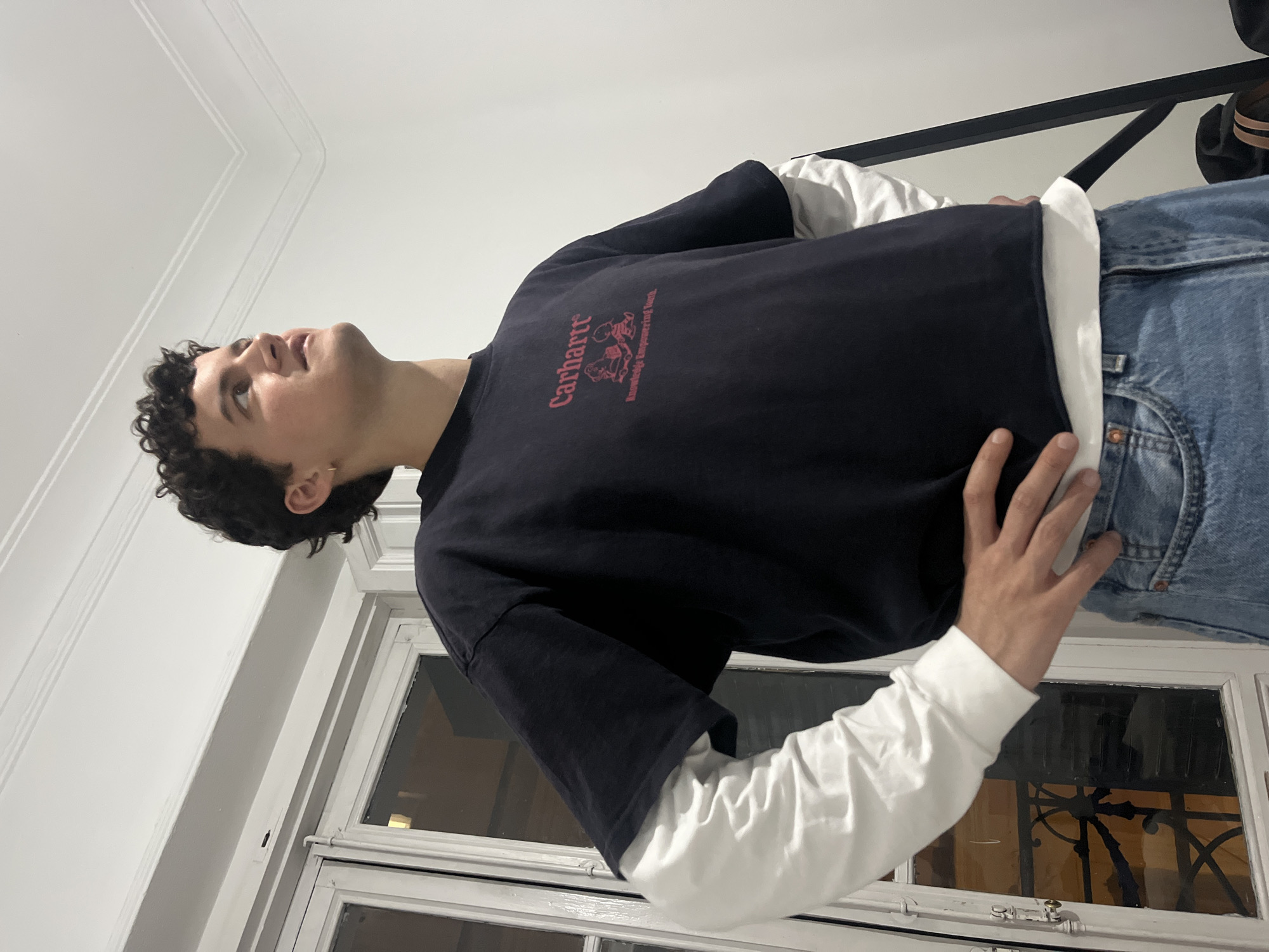 A young man with curly dark hair is looking upward and to the left in an indoor setting. He is wearing a long-sleeved white shirt under a black Carhartt t-shirt with red graphics. He is wearing blue jeans. He stands in front of a white window with a dark, ornamental design visible through the glass. The wall is white. The image orientation is vertical, with the person rotated slightly counter-clockwise. Without further information, it is impossible to determine the precise location within Madrid, Spain.