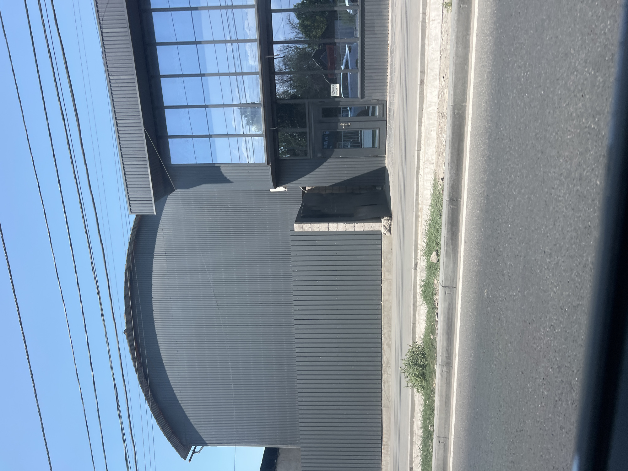 The image captures a street view in Bishkek City, Kyrgyzstan. In the foreground, a portion of a paved road is visible, with a concrete curb and a narrow strip of dry grass and weeds to its right. Behind the curb, a grey corrugated metal fence runs horizontally across the frame. Adjacent to the fence, a modern building with a grey corrugated metal facade and large glass windows stands. The building has a section with a curved, arched roof and another section with a flat roof and a prominent overhang above the windows. The windows reflect the blue sky and trees. A glass-paneled door is visible on the ground floor of the building, and a sign with Cyrillic text is mounted above it. Overhead, multiple power lines stretch across the clear blue sky. The overall scene suggests an urban industrial or commercial area.