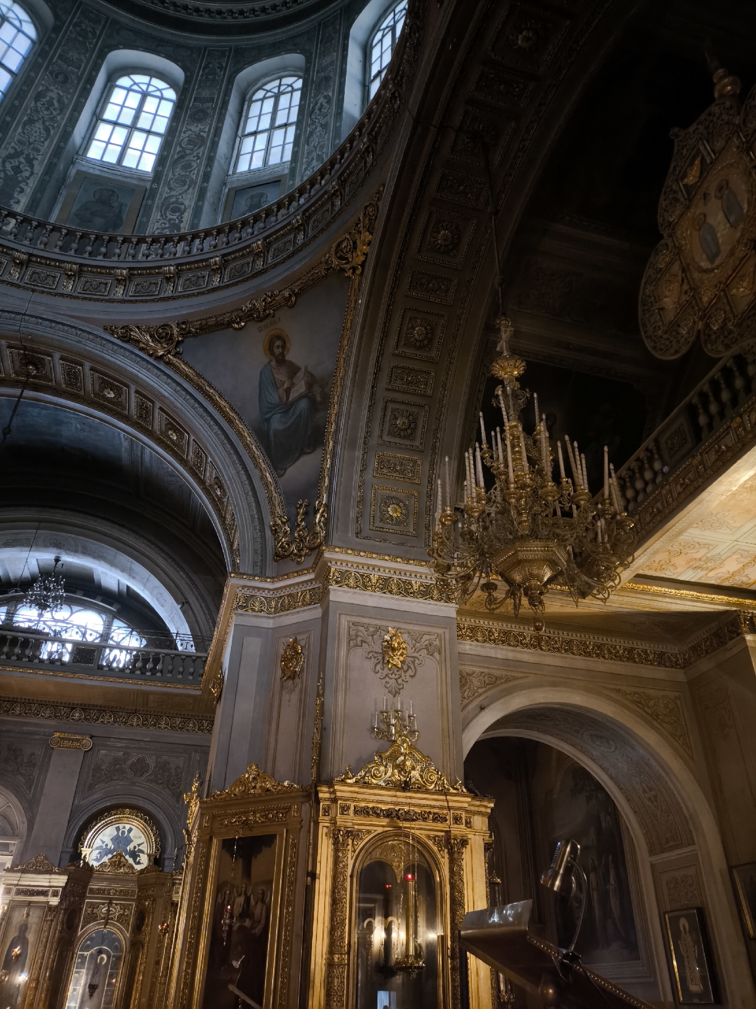 This is an interior view of a richly decorated Orthodox church, likely in an Eastern European or Russian city. The location is indicated as Unknown, Unknown.
**Architecture:**
The image showcases intricate architectural elements, including soaring arches, ornate columns, and a large dome. The walls are adorned with detailed moldings, gilded accents, and religious iconography. Several windows are visible near the dome.
**Objects:**
The scene contains a prominent chandelier with multiple arms and candles. There's also a lectern with a reading lamp, and portions of the iconostasis (altar screen) are visible. The iconostasis features gold-framed icons.
**Art & Decoration:**
Murals or paintings, likely of religious figures, decorate the walls. There are also various smaller gilded ornaments and wall decorations.
**Environment:**
The lighting appears to be a combination of natural light from the windows and artificial light from the chandeliers and sconces. The overall atmosphere suggests a grand and sacred space.