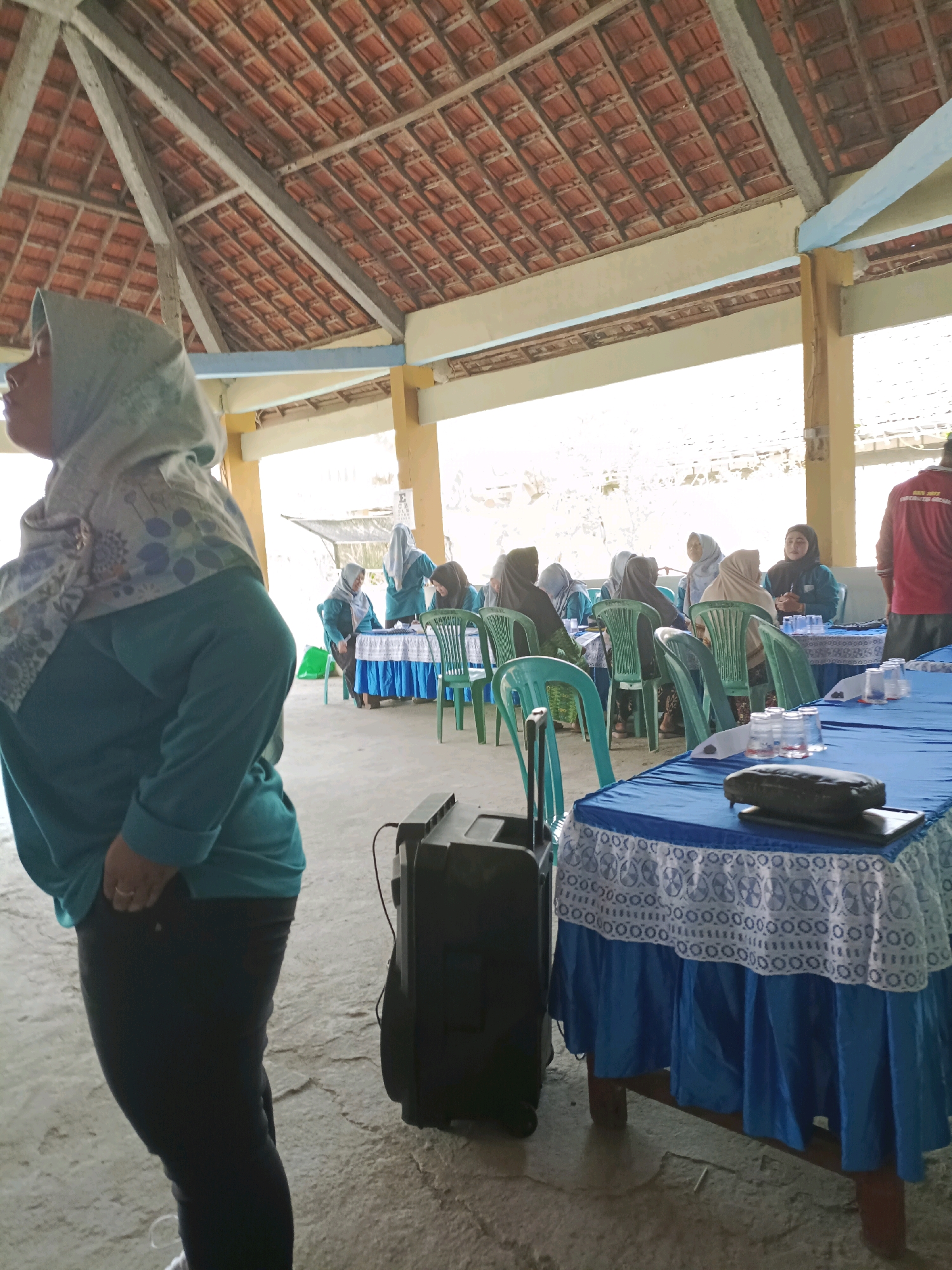 The image depicts an outdoor gathering under a covered structure with a tiled roof and wooden beams. In the foreground, a person wearing a teal hijab and matching long-sleeved shirt and dark pants stands facing away from the camera. To their left, a black speaker is visible. In the background, several individuals, mostly women wearing hijabs, are seated at tables covered with blue tablecloths. Some are interacting with each other, and one appears to be speaking. Green plastic chairs are arranged around the tables. The ground is concrete. The setting appears to be a community event or meeting in Rancang Kencono, Indonesia.