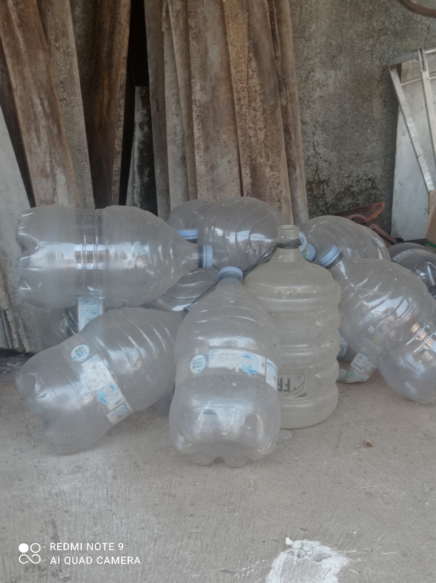 A group of empty, clear plastic water bottles are piled on a concrete floor in front of a textured, grey wall and a stack of weathered, vertical wooden planks. A metal ladder is visible leaning against the wall to the right. Some of the bottles have visible labels. The scene appears to be outdoors or in an open structure. The image is of a scene in Cipawon, Indonesia.