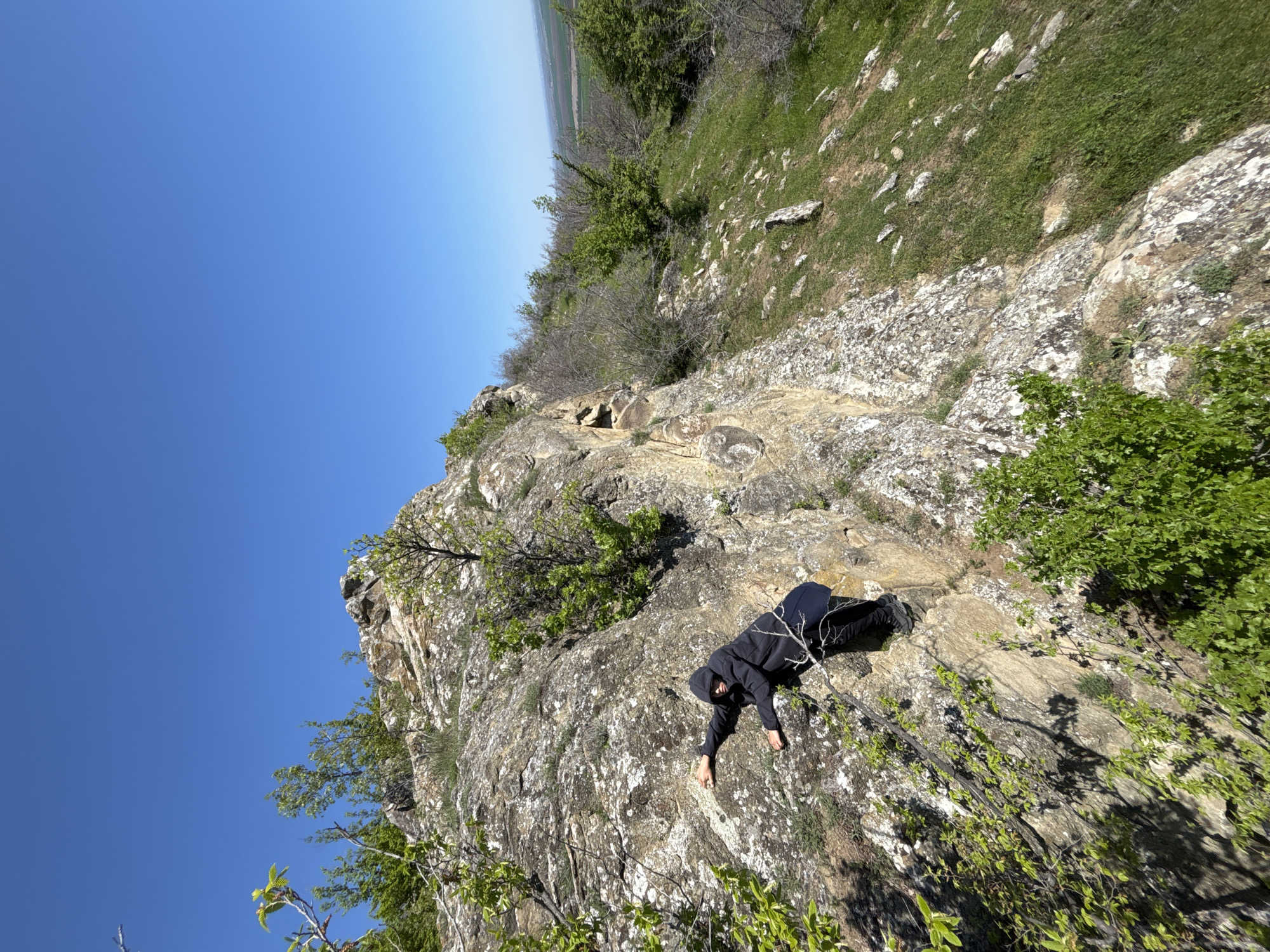 The image depicts a person in a dark hooded sweatshirt and dark pants lying prone on a rocky cliff face, likely in the vicinity of Goritsa, Bulgaria given the request for location. The person is positioned diagonally across the frame with one arm extended down the cliff. The cliff consists of light-colored rock with patches of moss and sparse vegetation. A clear blue sky occupies a significant portion of the upper left corner of the image. The upper right of the image shows a grassy hillside with some trees and shrubs. The photograph is taken from an elevated perspective, looking down on the cliff and the person.