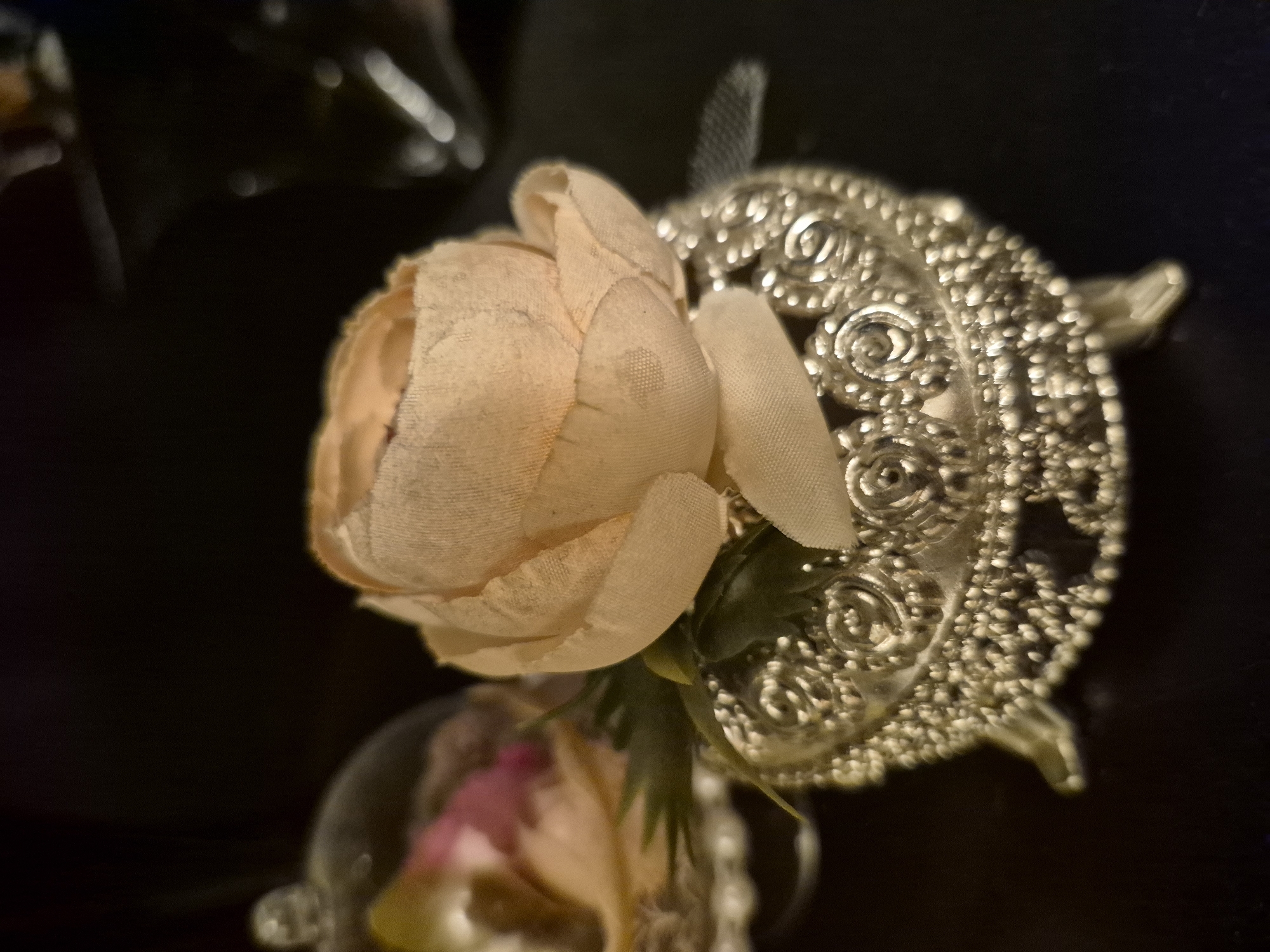 A close-up, shallow depth-of-field, dimly lit, indoor still life photograph, with a focus on a decorative silver trinket box topped with a light peach fabric flower. The box is ornate, with intricate scrollwork and a beaded edge, supported by four small, decorative legs. Green fabric leaves emerge from behind the flower. To the left and slightly out of focus, another decorative item is visible under a glass dome, containing pink and yellow fabric elements. The background is dark and blurred, with faint bokeh highlights suggesting other objects or light sources. The scene is forensic in its detail, capturing the textures of the fabric flower and the metallic sheen of the box. No people or actions are depicted. The setting is not identifiable as being in Vanadzor, Armenia, or any specific city.