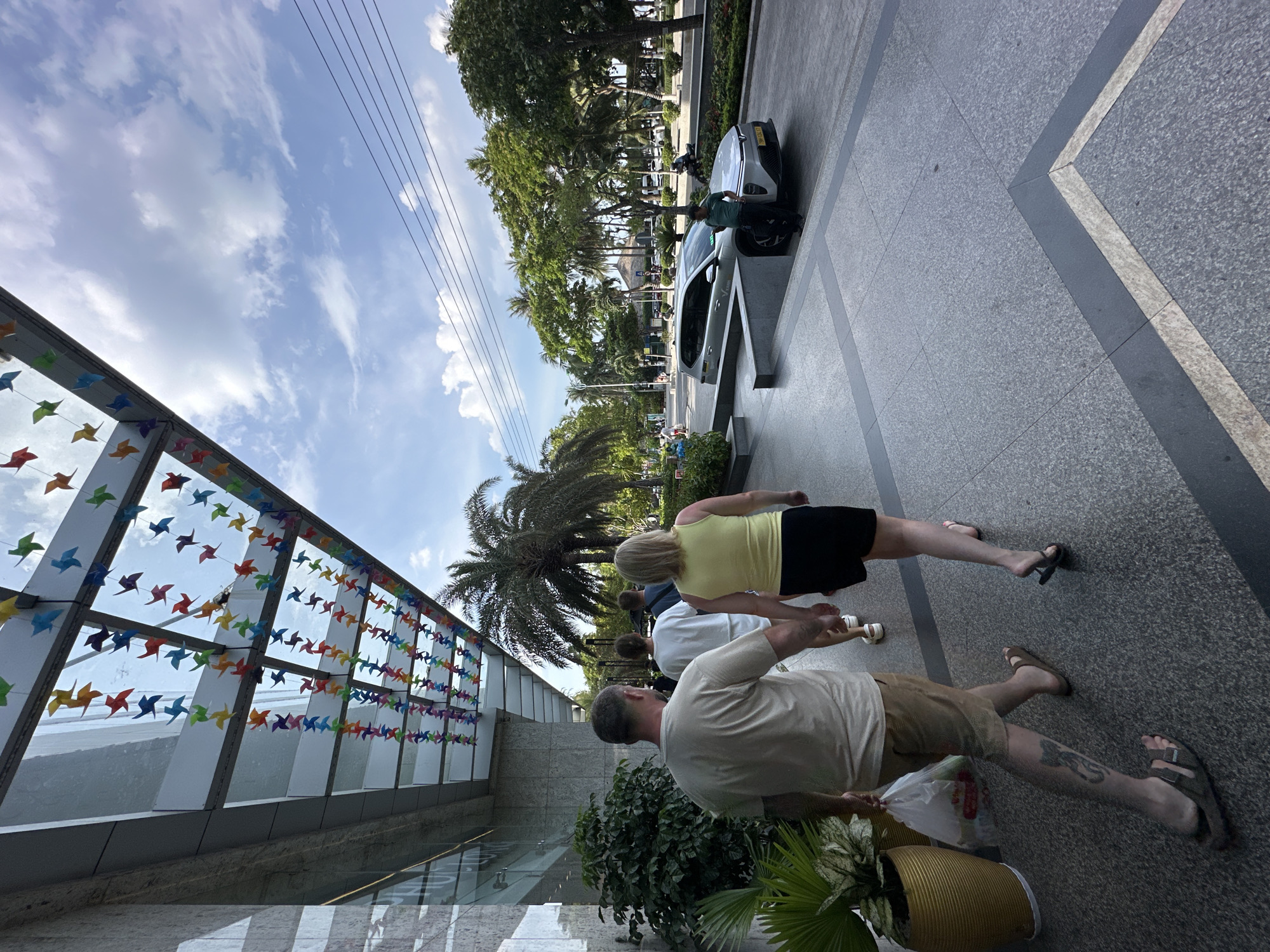 A group of people walk along a paved sidewalk next to a modern building. The building has a glass facade adorned with strings of colorful pinwheels in various shades of blue, green, yellow, red, and purple. Overhead, power lines stretch across a bright blue sky dotted with fluffy white clouds. Lush green trees, including palm trees, line the street, suggesting a tropical or subtropical climate. A white car is parked on the street next to the building, with a man leaning over the open driver's side door.
The people are walking away from the camera, mostly in profile. The foremost individuals are a man wearing a light-colored t-shirt and khaki shorts, and a woman in a yellow tank top and black shorts. Both are wearing sandals. Behind them, another person in a white t-shirt is visible, and further back, another individual. The overall scene suggests a casual daytime activity, possibly sightseeing or walking through a commercial area. The sunlight and clear sky indicate pleasant weather. The presence of palm trees and the mention of Nha Trang, Vietnam, further reinforce the tropical setting. No visible text is present in the image.