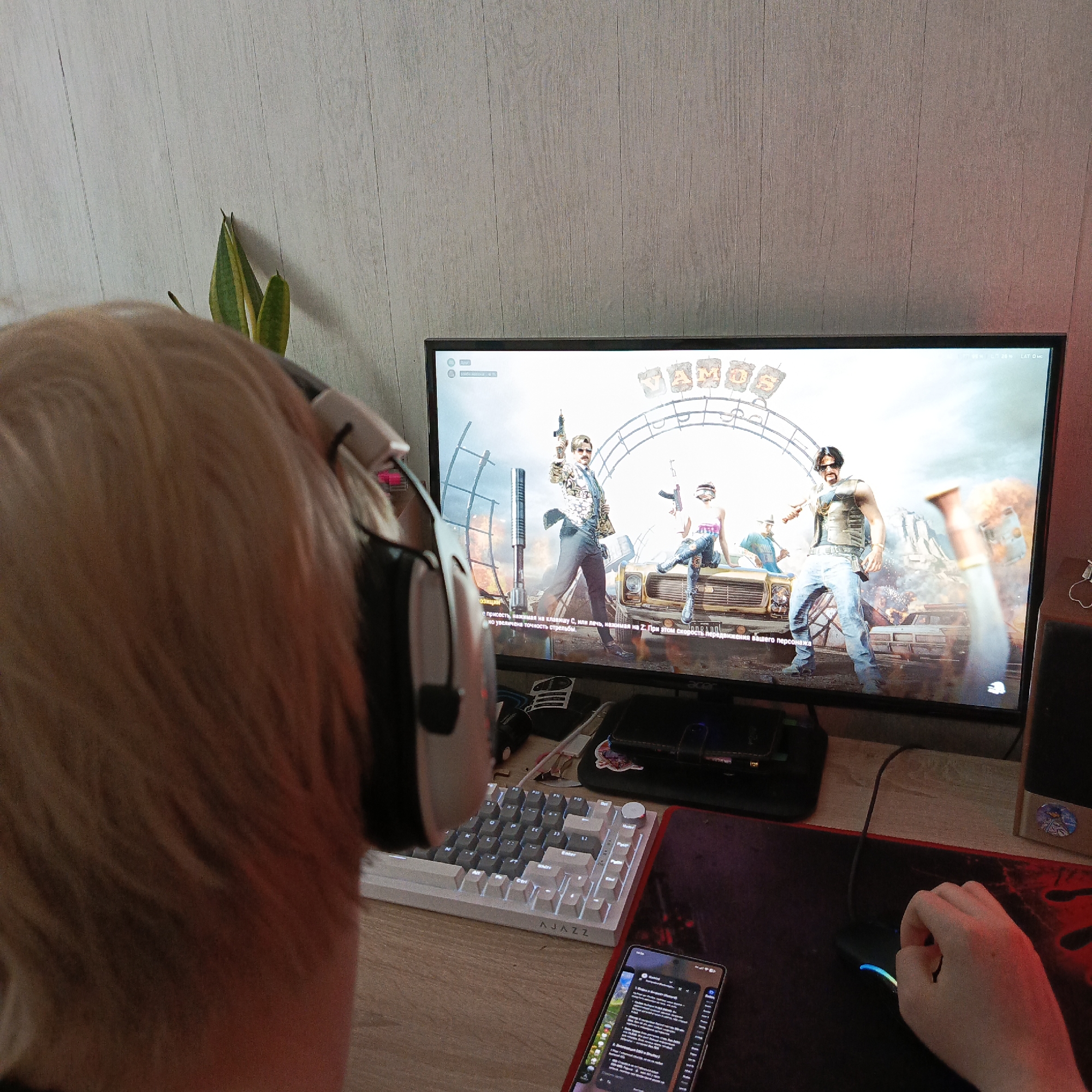 A person wearing headphones is sitting at a computer desk, playing a video game. The game displayed on the monitor appears to be an action or shooter title, featuring several characters and a stylized backdrop. A white keyboard with the brand name "AJAZZ" is visible on the desk, along with a gaming mouse and a mousepad with a red design. A smartphone with a screen displaying text is also present on the desk, near the keyboard. A plant stands behind the monitor. The location is not indicated in the image.