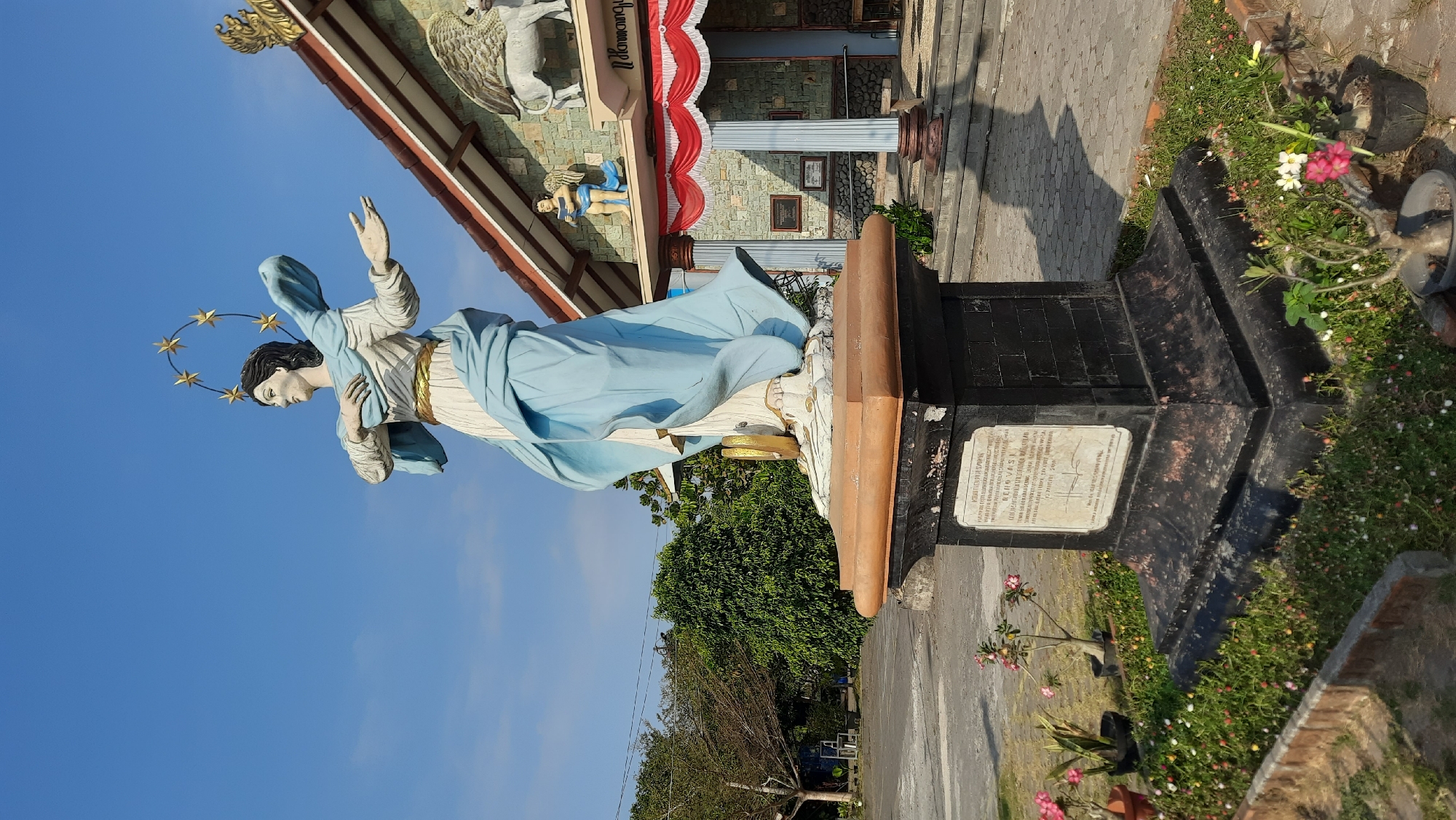 A full-shot, eye-level outdoor daytime photograph captures a statue of the Virgin Mary in front of a building in Nanggulan, Indonesia. The statue, adorned with a golden halo of stars, is dressed in a blue robe with a golden belt and a white undergarment. It stands on a dark, multi-tiered pedestal with an inscription plaque on its front. The building behind the statue features a stone facade, a red and white draped awning, and decorative elements on its roof, including a winged bull sculpture. Green trees and a clear blue sky form the background. The foreground is decorated with small flowering plants in pots and a brick border.