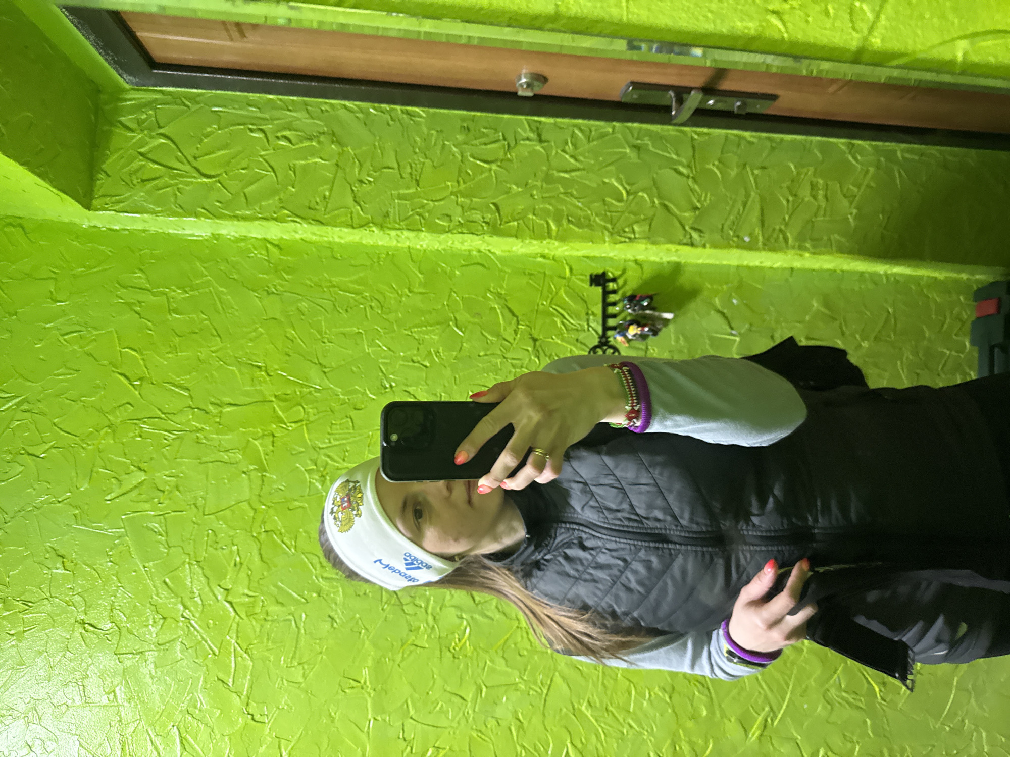 An indoor mirror selfie captures a female individual holding a black smartphone. The image is oriented with the person lying on their side, indicating a rotation from a vertical mirror reflection. The woman stands in front of a strikingly textured, bright lime green wall. Above her reflection, a wooden door with a dark frame and silver hardware is visible, likely a handle or lock, suggesting an entryway or hallway. To the right of her head, partially obscured, is a wall-mounted key organizer holding several keys.
The woman is dressed in a light grey long-sleeved top under a black quilted vest. She wears a white headband or buff featuring a design resembling the Russian double-headed eagle, with the word "РОССИЯ" (Russia) above it. Below the emblem, the word "adidas" is visible, mirrored to appear as "adepag" due to the reflection. Her nails are painted a vibrant orange or coral color. She accessorizes with multiple bracelets, including purple and multi-colored beaded ones, on both wrists, and rings on her left hand. Her long brown hair flows around her shoulders. The setting, characterized by the distinctive green wall, door, and key holder, indicates an indoor environment, likely a home in Chaikovsky, Russia.