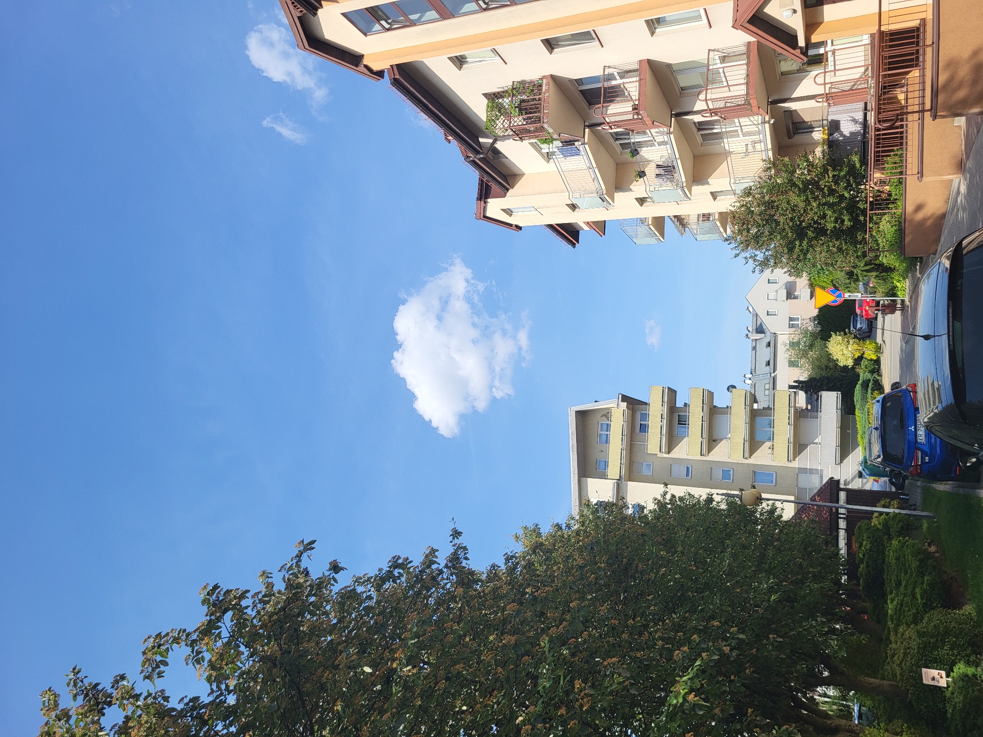 The image shows a view of buildings and foliage under a blue sky with a few clouds. The buildings are multi-story, with balconies and a tan color scheme. The foliage includes trees with green leaves. A street with cars is visible between the buildings. The image is taken from a slightly low angle, looking upwards.