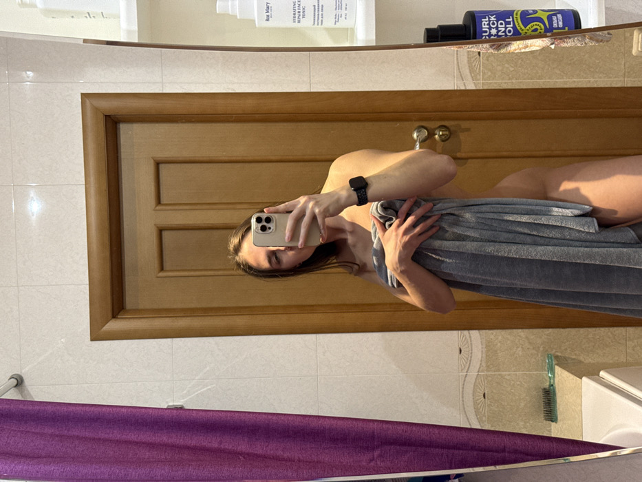 In a mirror selfie taken in a bathroom, a person covers themselves with a large grey towel. The person is wearing a smartwatch on their left wrist and is holding a smartphone to take the picture. A wooden door is reflected behind the person, with bathroom essentials visible on a shelf above. The lower part of the reflection shows a purple cloth and bathroom tiles. The scene is generally lit, suggesting a bright environment.