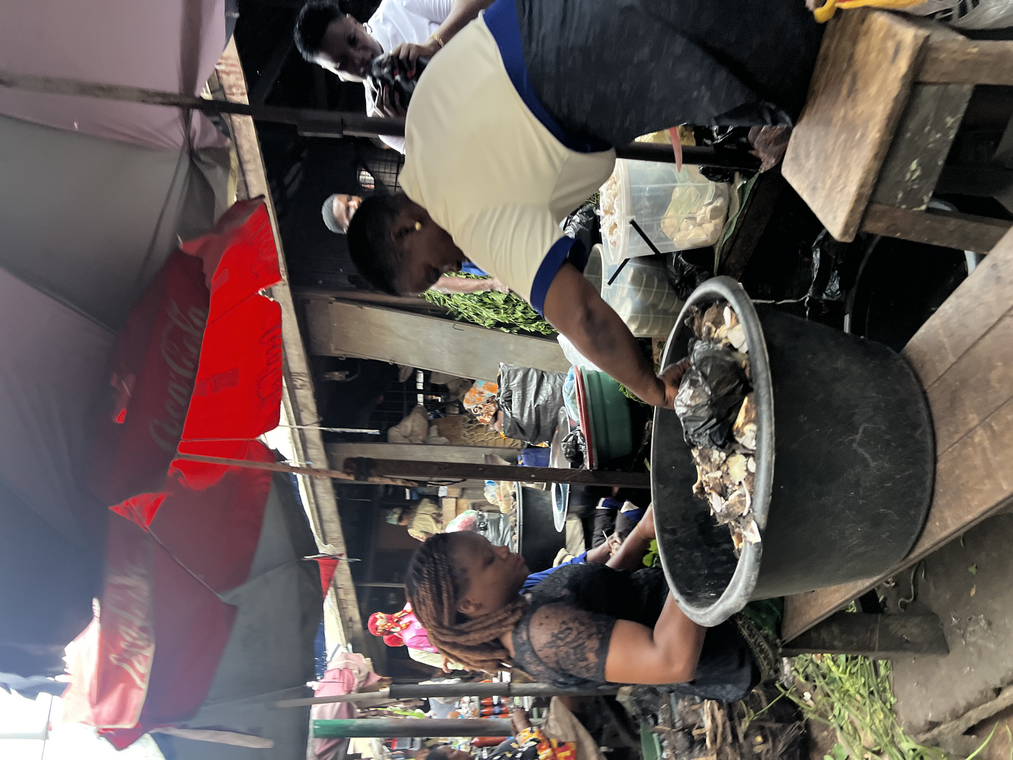 This image depicts a bustling market scene in Nkpor, Nigeria, likely a section dedicated to food vendors. In the foreground, a woman with braided hair wearing a black lace top and a green patterned skirt is actively engaged in her work, tending to a large black tub filled with what appears to be processed food items, possibly smoked fish or cassava products. To her right, another woman, wearing a pale yellow and blue top, is also focused on her task, reaching into a clear plastic container filled with similar food items. Both women appear to be vendors, serving customers or preparing goods for sale.
Behind them, the market environment is visible, characterized by makeshift stalls and awnings. Red and grey umbrellas provide shade, one of which prominently displays a Coca-Cola logo. Other market-goers can be seen in the background, some obscured by the foreground subjects. The ground appears wet and somewhat uneven, suggesting it's an outdoor market. The overall atmosphere is one of lively commerce, with the women at the center of the activity, showcasing the typical hustle and bustle of Nigerian markets. The presence of diverse food items, the interaction between vendors, and the crowded setting all point to a vibrant marketplace where daily transactions and livelihoods unfold.