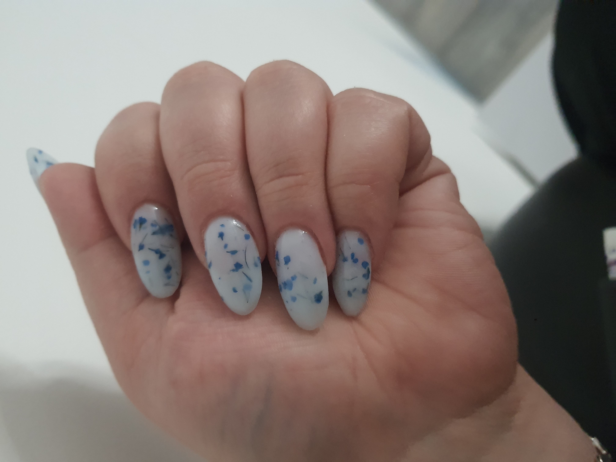The media file shows a close-up of a person's hand with manicured nails. The nails are a light, translucent white with small, scattered blue flecks, giving them a delicate, natural appearance, possibly resembling pressed flowers or confetti. The hand is resting on a white surface, which appears to be a table or countertop. The lighting suggests an indoor setting, possibly a well-lit room. The background is blurred, but hints of a darker object and a light, possibly grey, wall or surface are visible. There is no visible text in the media file. The overall impression is one of personal grooming and aesthetic presentation.