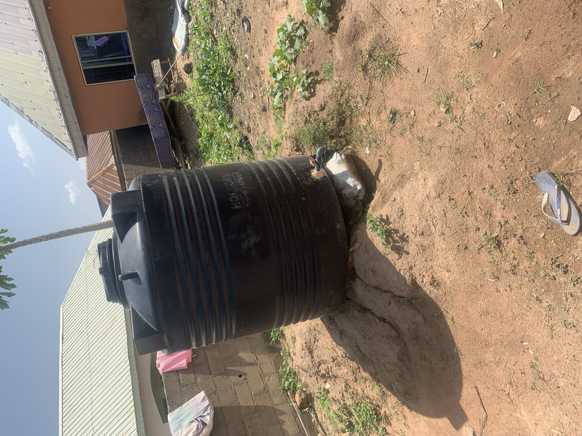This media file shows an outdoor scene on a sunny day, likely in Jalingo, Nigeria. The central object is a large black water tank, supported by a concrete structure. A pipe with a red valve is attached to the top of the tank, suggesting it is either being filled or water is about to be dispensed.
In the background, there is a building with a light brown facade and a window. To the left of the building, a portion of a house with a light-colored corrugated metal roof is visible. There are also some construction materials or debris, including a blue and gold mattress leaning against the building and a pile of what appear to be bricks.
The ground is predominantly dirt with patches of sparse green vegetation. A pair of blue flip-flops is visible on the right side of the image, lying on the sandy ground. A tree with green leaves is partially visible on the far left, its trunk extending upwards and slightly into the frame. The overall impression is of a functional outdoor space, possibly a residential or community area, with emphasis on water storage. The bright sunlight and clear sky indicate it's daytime, and the scene appears to be relatively calm, with no people actively engaged in any activity. Some faint text is visible on the water tank, reading "HOLBORN QUALITY WATER TANKS".
