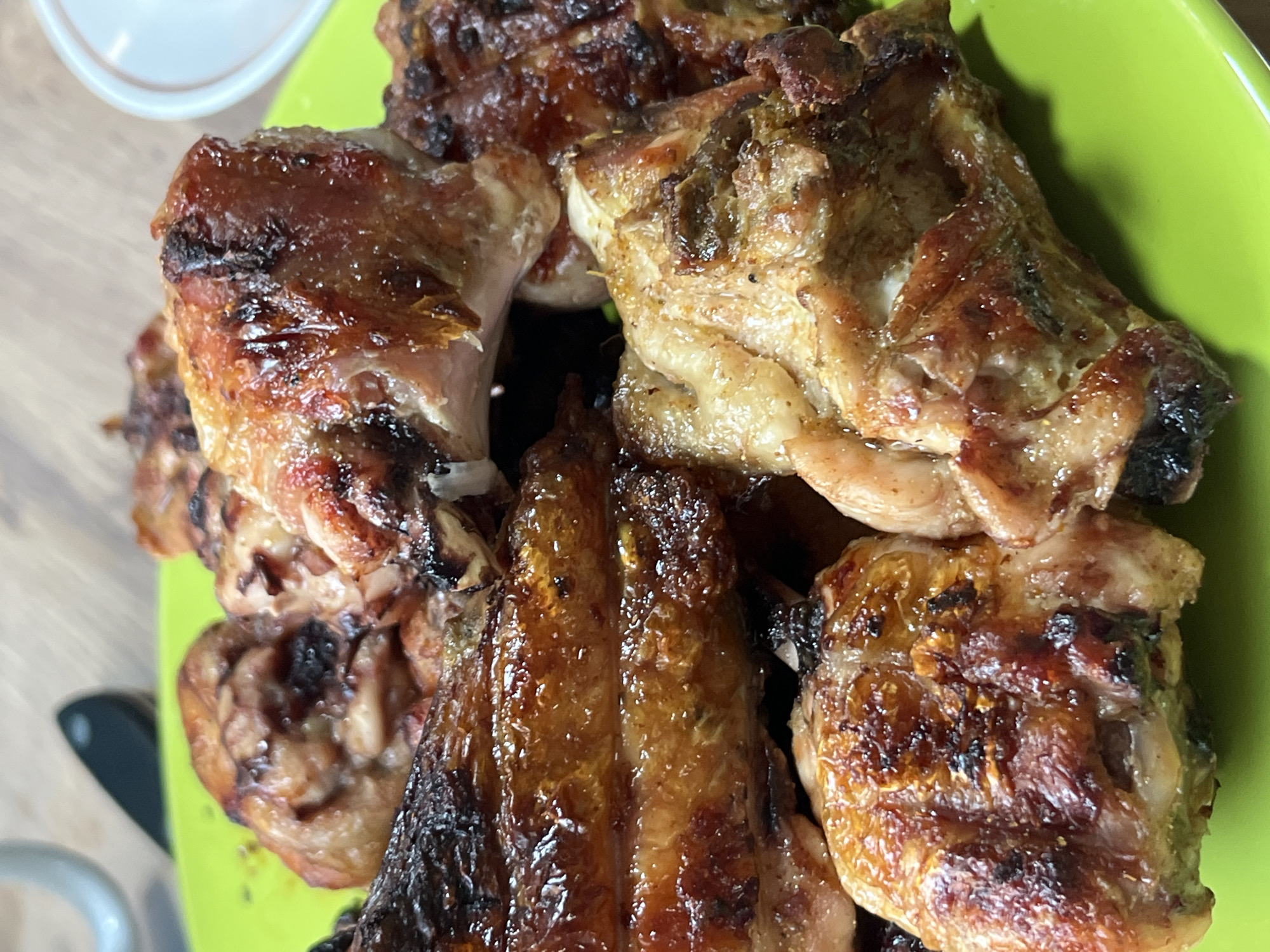 The image shows grilled chicken pieces piled on a green plate. The chicken appears to be well-cooked, with visible grill marks and a glossy surface. There is a light-colored object, possibly a small bowl, visible in the upper left corner. The background is a wooden surface. The location is not discernible from the image alone.